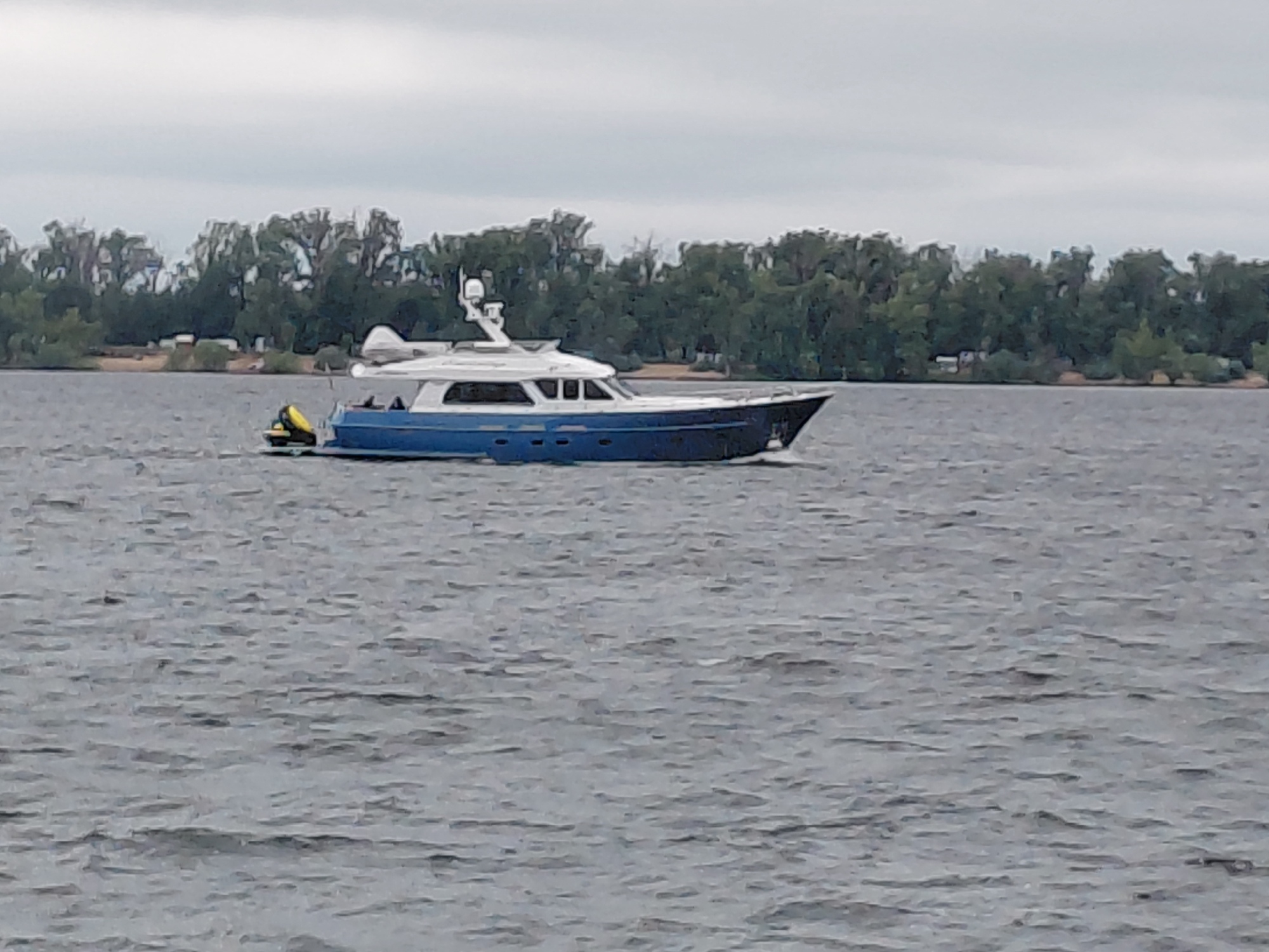 A blue and white motor yacht is cruising on a grey, choppy body of water. The yacht has a sleek design with a white superstructure and a blue hull. It is moving from left to right in the frame. In the background, there is a dense line of green trees bordering the water, suggesting a natural landscape. The sky above is overcast and grey, contributing to the muted color palette of the scene. There are no people visible on the yacht or in the surrounding environment. The location is the Volga River near Novokuybyshevsk, Russia.