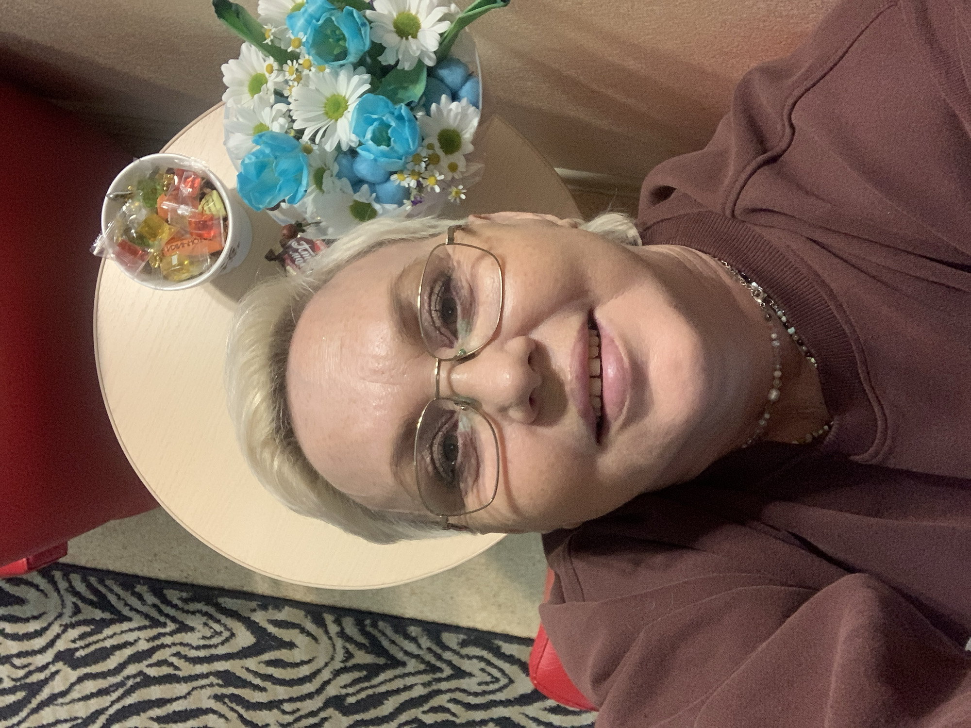 A high-angle selfie depicts an adult female with short, blonde hair and gold-rimmed rectangular eyeglasses, smiling directly at the camera. She is wearing a dark reddish-brown sweatshirt and a multi-strand necklace. She appears seated in a red chair, portions of which are visible on the left and right. To her upper right, a light-colored, circular table holds a small white cup filled with individually wrapped candies and a bouquet. The bouquet consists primarily of white daisies, blue tulips, and small white filler flowers, with some small blue decorative objects intermingled. The immediate background to the left features a carpet with a black and tan zebra print pattern. Behind the table, a light brown wall or surface is visible. This scene is located in Chaikovsky, Russia.