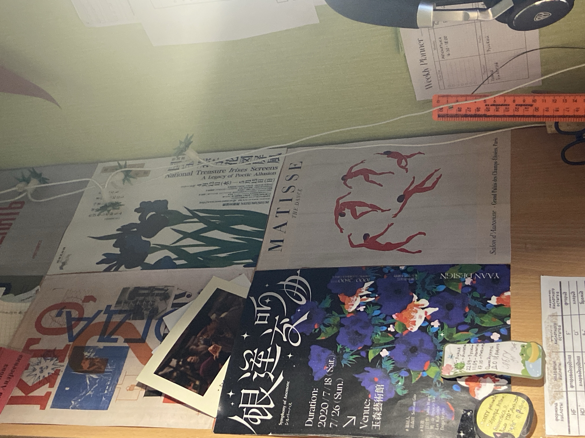 This image captures a close-up, overhead view of a desk cluttered with various papers, books, and art prints. The setting appears to be a creative workspace, possibly a home office or studio.
The dominant subjects are art prints, including a reproduction of Henri Matisse's "The Dance," a print with figures in red, and another featuring colorful floral motifs and koi fish. There are also what appear to be exhibition posters or flyers, one with Japanese text and imagery of irises, and another with bold graphic lettering. Mixed in are what look like personal photographs or small prints, and a "Weekly Planner" with entries in Russian, indicating a scheduled activity or appointment.
Visible text includes titles of artworks like "National Treasure Irises Screens," "MATISSE THE DANCE," and "Symphony of Anemone." There's also text in English and Russian, including dates like "2020/7/18 (Sat.)" and "7/26 (Sun.)," and a ruler marked with centimeters. A pair of headphones is partially visible at the top of the frame, suggesting the presence of music or audio equipment.
The overall impression is one of creative output and organization, with artistic elements juxtaposed with practical planning tools. The lighting is somewhat dim, suggesting an indoor setting. No specific time of day or weather is discernible. The presence of Russian text, coupled with the location context, indicates the setting is Zavodoukovsk, Russia.