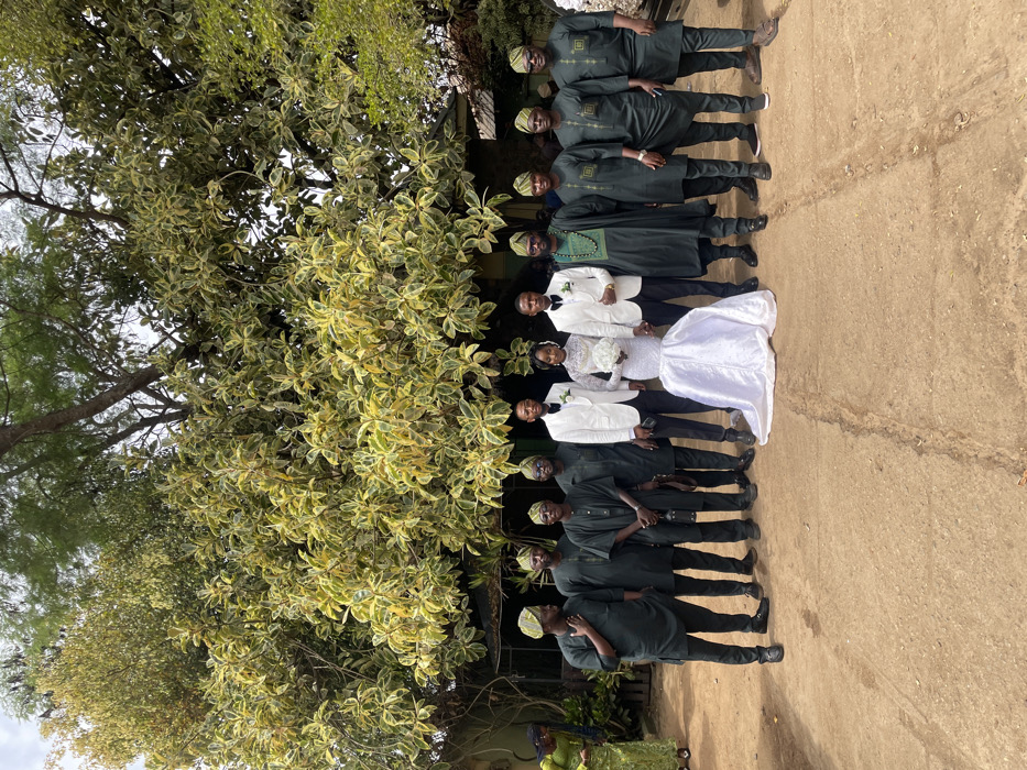 A group of twelve individuals, including a bride and groom, are formally posed outdoors for a photograph. The central bride wears a white, long-sleeved, high-necked mermaid-style wedding gown with a white floral bouquet. The groom, to her immediate right, is dressed in a white tuxedo jacket, black trousers, white shirt, and black bow tie. Two additional men, positioned one to the bride's left and one to the groom's right, wear identical white tuxedos. Eight other men, flanking the central party, are attired in dark green or black traditional Nigerian outfits, comprising tunics, trousers, and matching caps (fila), some featuring subtle embroidery. All primary subjects face the camera. The ground is reddish-brown compacted earth. The background features a large tree with variegated green and yellow leaves, and partially visible building structures with a light-colored wall and dark roof. A woman in a green patterned dress is partially visible on the far left, bending down. The specific location in Jos, Nigeria, is not identifiable from the visual content; however, the attire and environment are consistent with a Nigerian outdoor event or residential setting.
