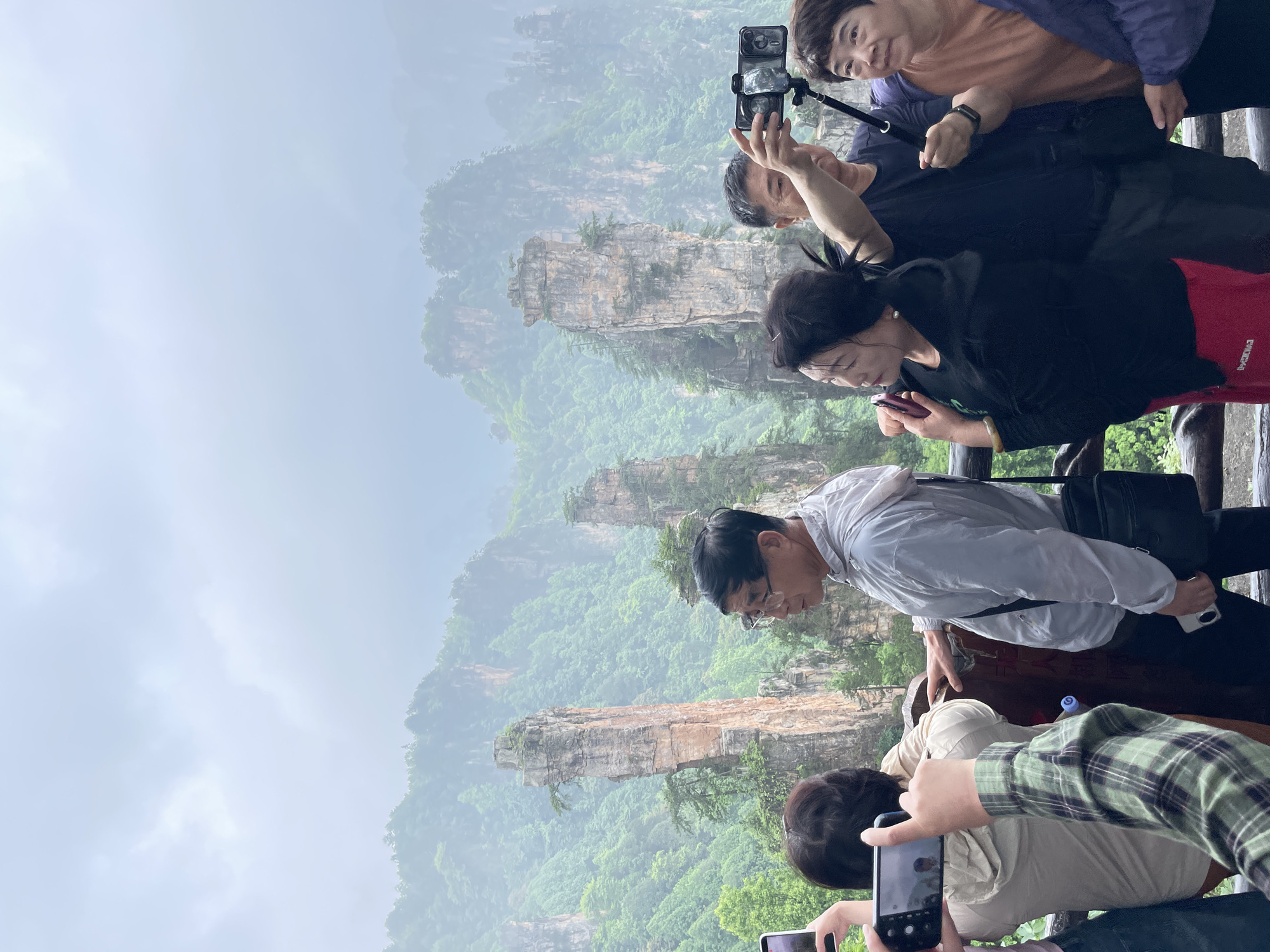 The image shows a group of people at a viewpoint in the city of Wulingyuan, China, likely in the Zhangjiajie National Forest Park. The park is known for its unique sandstone pillars. A layer of fog hangs in the air.
The people are standing near a dark wooden railing. They appear to be tourists, with several holding smartphones and taking pictures of the landscape. Some are looking at their phone screens.
The landscape features numerous tall, slender rock formations covered in greenery. The sky is overcast, suggesting a cloudy day. The overall scene depicts a tourist setting in a scenic natural area.