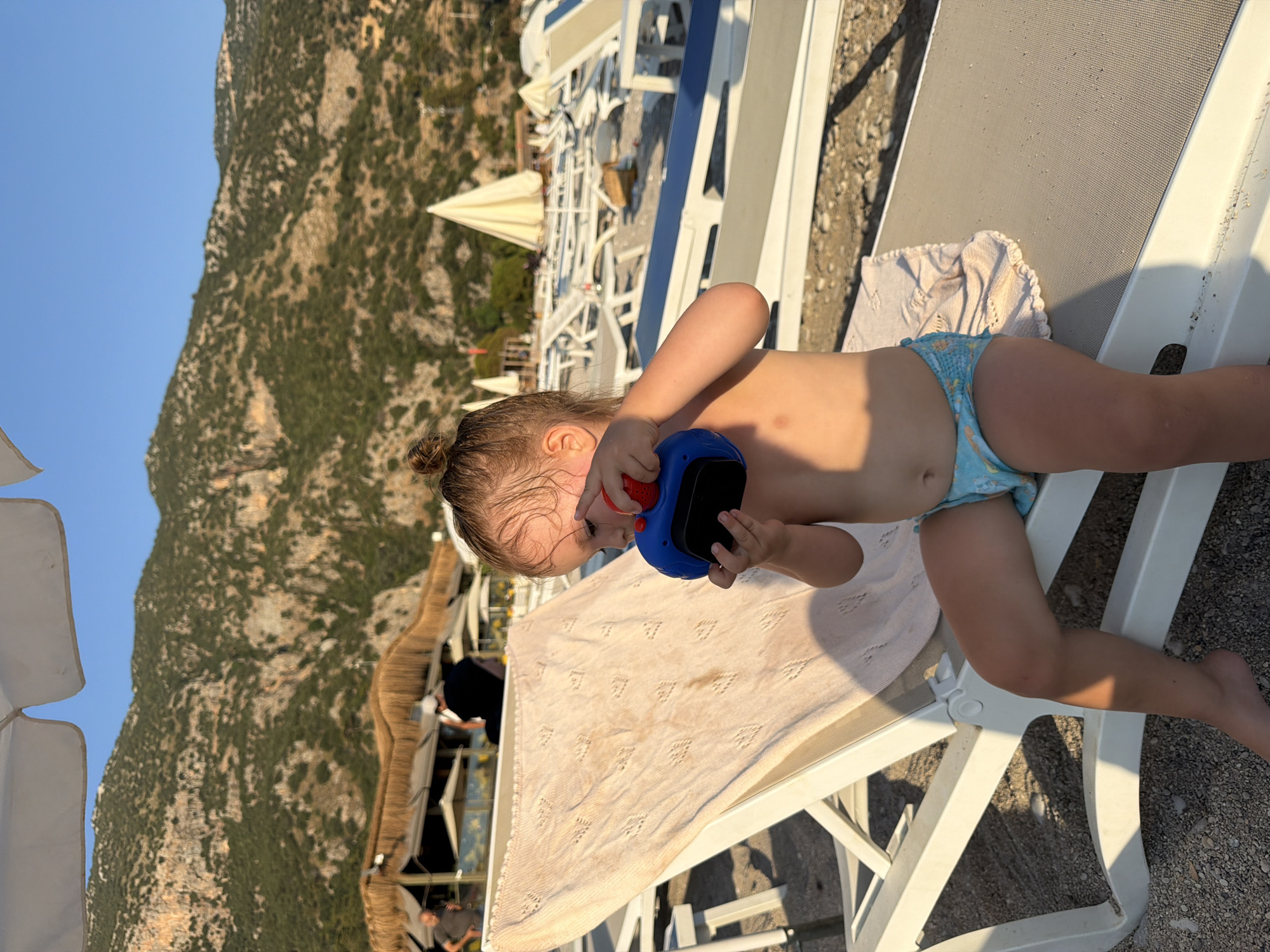 The image shows a young child lying on a sun lounger at a beach. The child is wearing blue patterned swim trunks and is holding a blue and red object, possibly a toy. The sun lounger has a beige towel draped over it. In the background, there are more sun loungers, umbrellas, and a mountainous landscape with green vegetation. The sky is clear and blue, suggesting a sunny day. The scene appears to be set in Fethiye, Turkey, indicated by the mountainous terrain and beach setting. The time of day seems to be late afternoon or early evening, as the sun is casting long shadows. The child's focus on the object in their hands suggests engagement and perhaps curiosity. The overall atmosphere is one of relaxation and enjoyment in a beautiful outdoor setting.