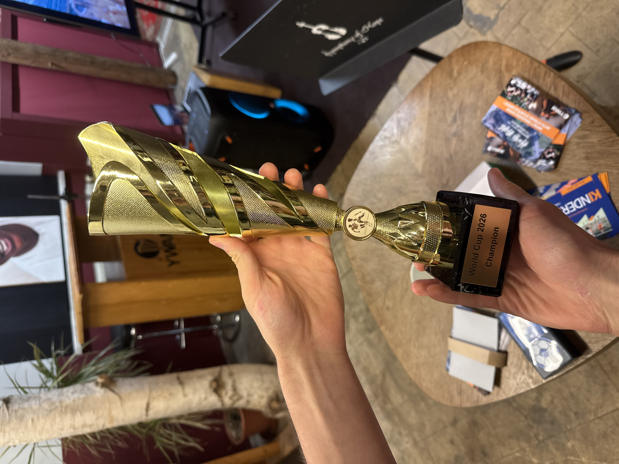 The image shows a close-up of two hands holding a gold trophy inscribed with "World Cup 2026 Champion". The trophy is placed on a small black marble base. In the background, a wooden podium with a logo that appears to be "YWA" is visible, along with a black speaker and a black screen displaying an image of a person. Other objects in the background include a potted plant, a tree trunk, and a wooden table. The scene appears to be indoors, possibly at an event or gathering in Bad Blankenburg, Germany.