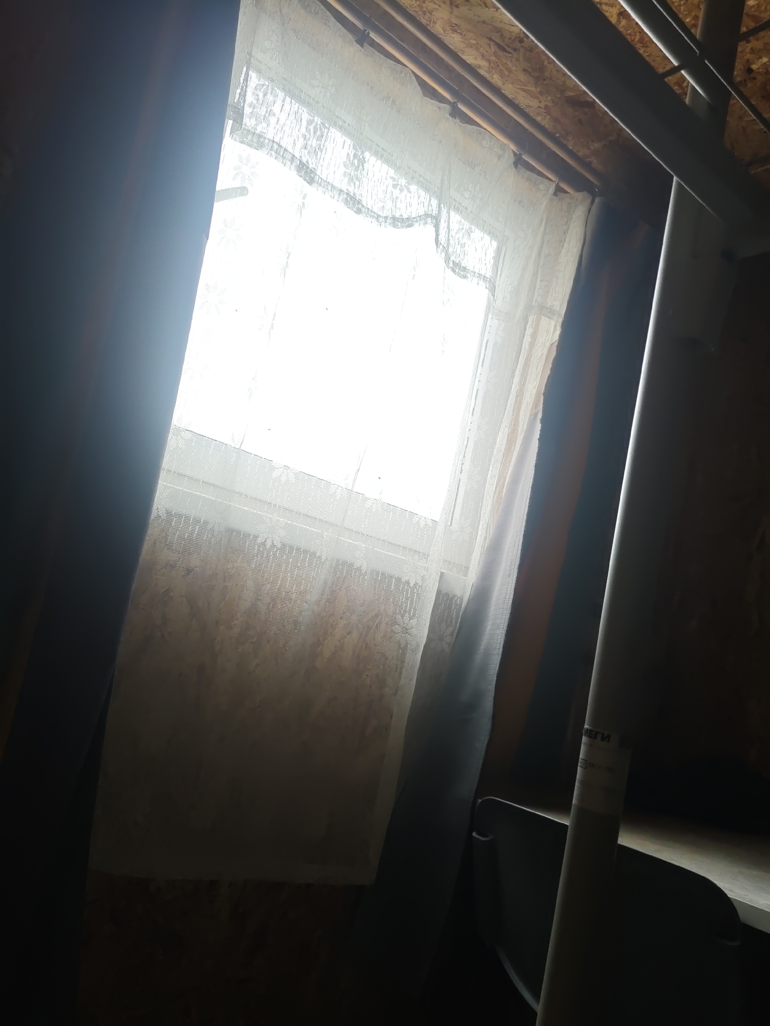 The image shows the interior of what appears to be an attic or unfinished room. A window is covered with a sheer white lace curtain, allowing bright daylight to filter through. To the left of the window, a dark curtain or fabric hangs down. To the right, a metal pipe or railing is visible, running diagonally upwards. The walls appear to be constructed of plywood or unfinished wood. On the metal pipe, there is a label with some text that is difficult to read, but appears to be in Cyrillic script. The overall impression is of a dimly lit, somewhat cluttered space with natural light coming from the window. The location is identified as the city of Платошино, Russia.