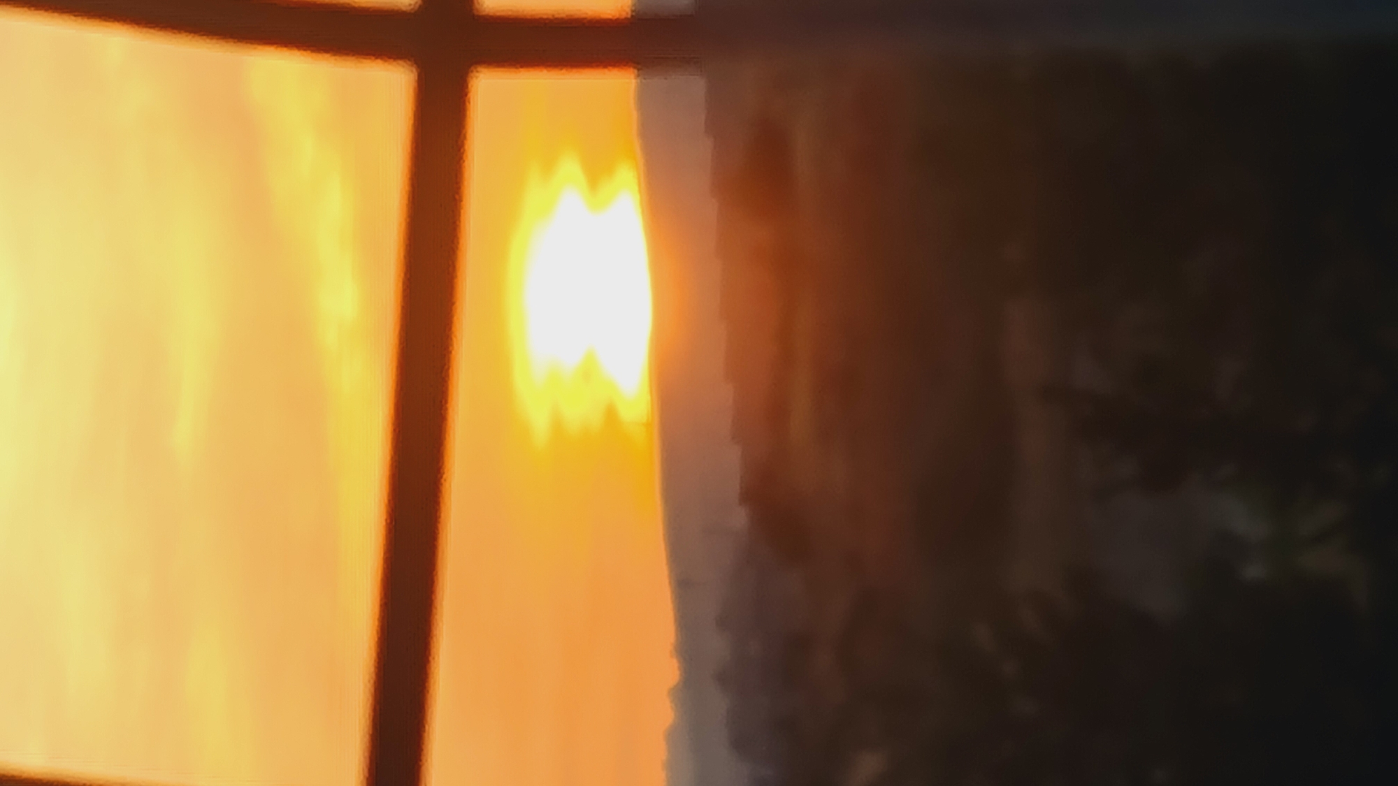 A blurry image shows a bright, orange sunset seen through a window with a dark frame. The area to the right of the sunset is dark and out of focus, possibly showing vegetation or a structure. The location might be Lachish Regional Council, Israel, but this is not visually discernible from the image.