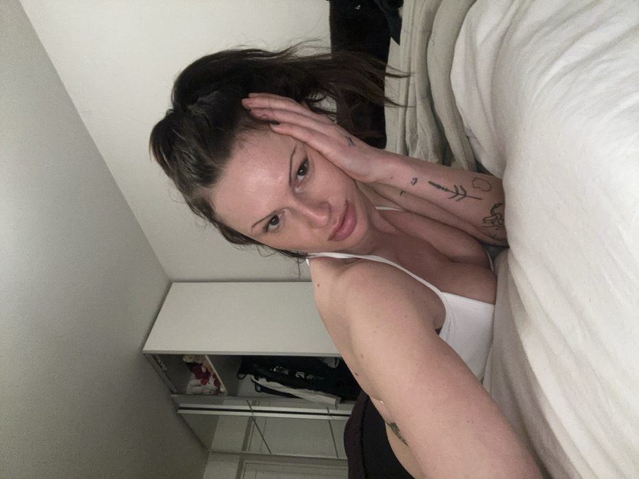 A fair-skinned woman with dark hair pulled back in a ponytail lies in a bed with white linens. She's wearing a white tank top. Her left arm is bent, her hand touching her forehead. Tattoos are visible on her left arm. A white storage unit with visible items is in the background. The camera angle is slightly askew, creating a tilted view.