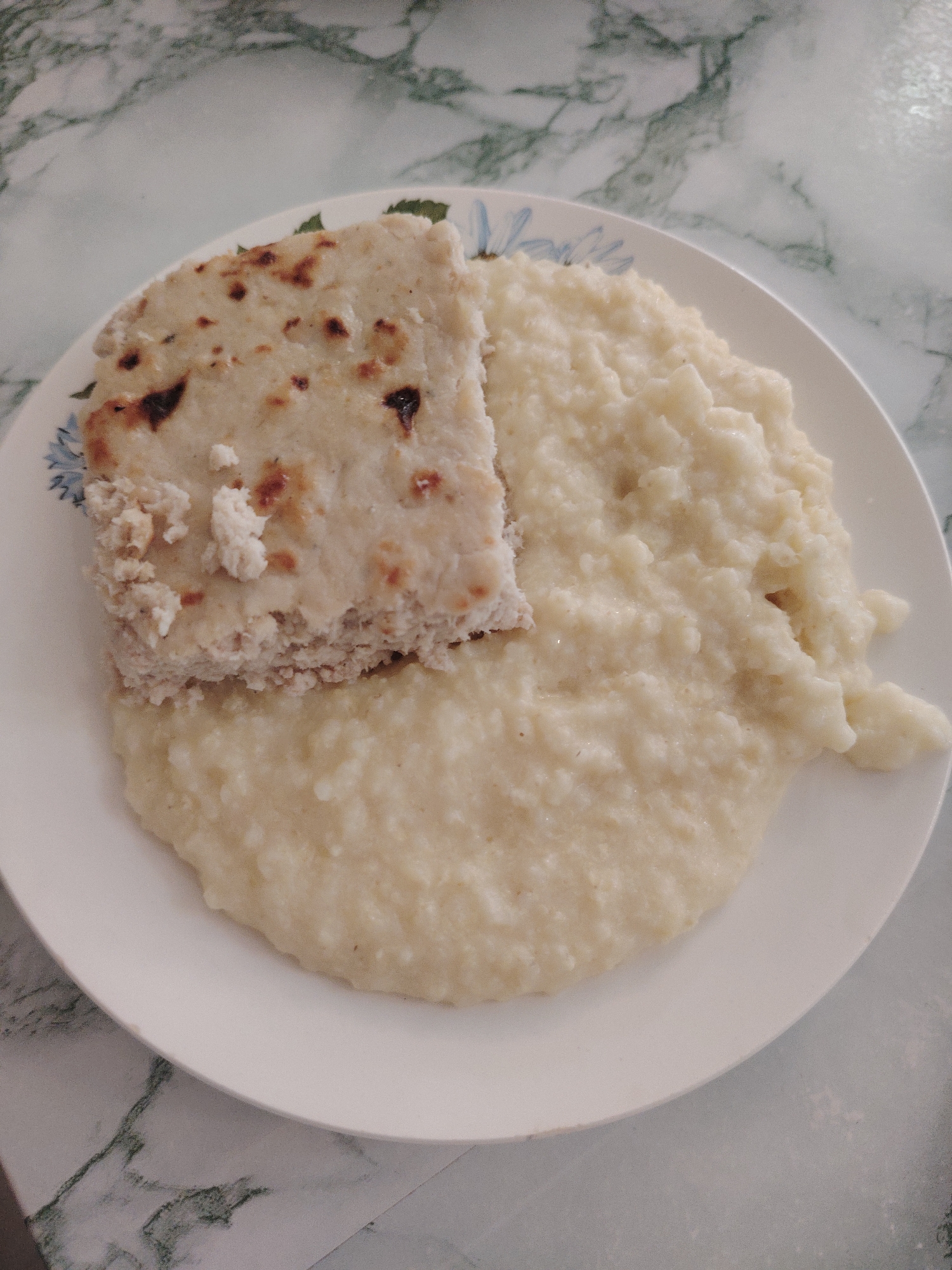 A plate of food is presented against a backdrop of a marbled surface. On the plate, there is a square piece of what appears to be flatbread or cake. Next to it is a portion of porridge. The plate has a floral design around the edge. The food is placed on a patterned surface that could be a table or countertop. There is no information about the city of Sarapul, Russia in the image.