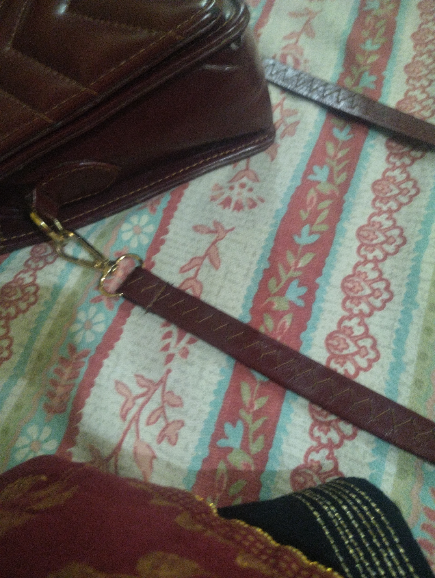 A close-up, overhead shot shows a brown quilted handbag with a dark brown leather strap attached to a gold clasp. The strap extends diagonally across a fabric surface with a repeating pattern of vertical stripes in white, red, and light blue, featuring floral and leaf motifs in pink and light blue. In the bottom left corner, a portion of a burgundy garment with gold embroidery is visible. The image is static and shows no activity. The location is Unknown, Unknown.