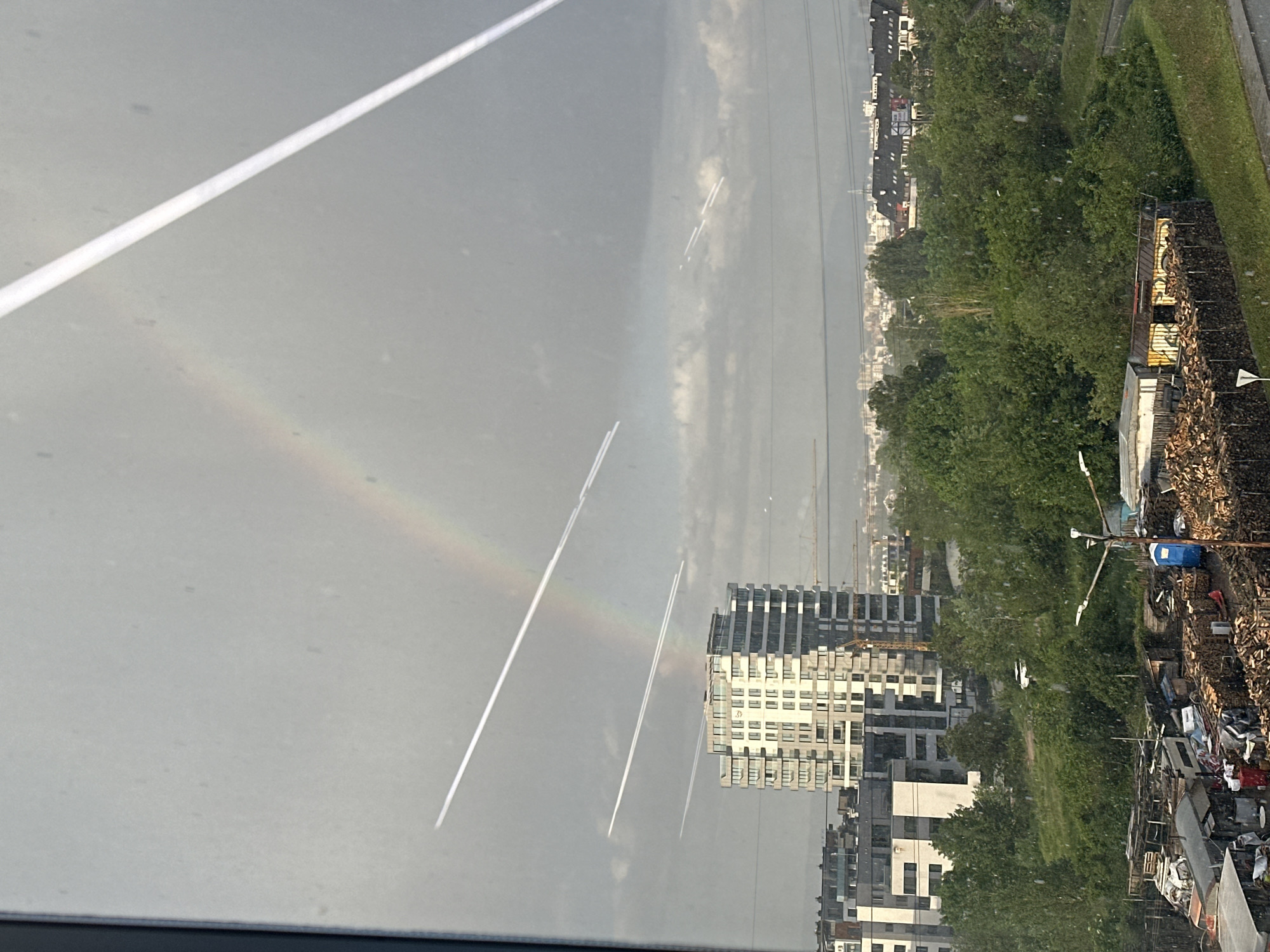 The image depicts a view of Sofia, Bulgaria, seemingly taken from inside a vehicle or building, looking out across an urban landscape. In the foreground, a road with a white line is visible. A faint rainbow stretches across the sky. Below the rainbow, there are airplane contrails. Modern, high-rise residential buildings are clustered in the middle ground, partially obscured by green trees. A construction crane can be seen near the buildings. Adjacent to the buildings and trees, a cluttered area with materials like wood and scrap metal is observed. The sky is overcast.