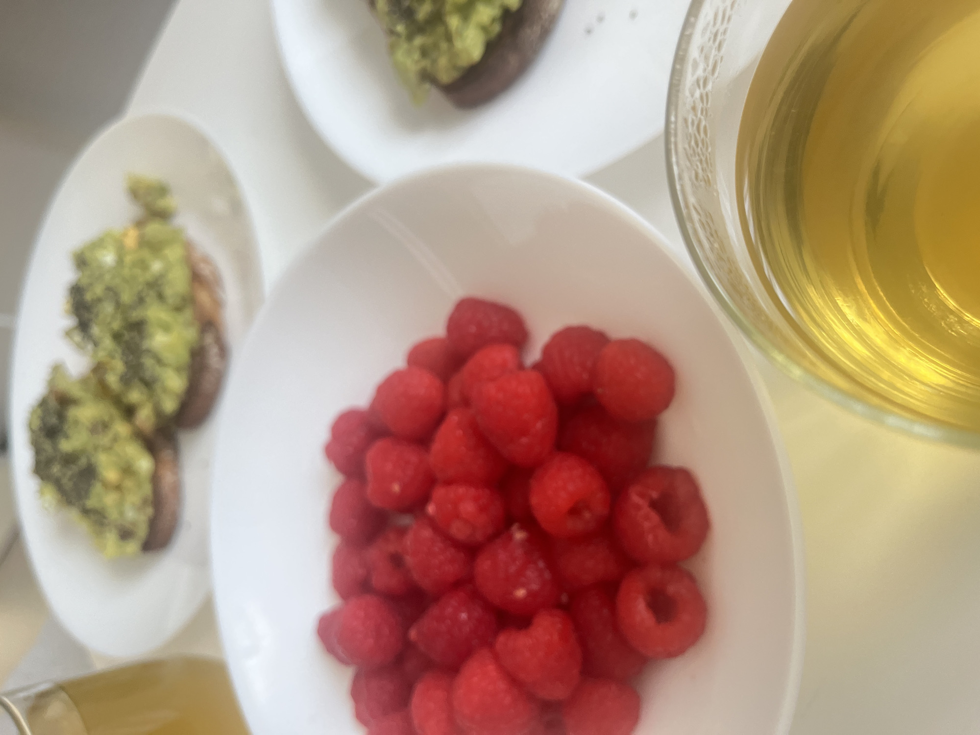 The image presents a close-up, overhead view of a light-colored surface featuring a meal consisting of raspberries in a white bowl, avocado toast on two separate white plates, and a glass of tea. The raspberries are bright red and clustered together in the center of the bowl. The avocado toast appears to be topped with a green spread. The tea in the glass is a light amber color. The scene seems to be a breakfast or light meal setting. While the city is specified, the exact location within Mogán, Spain is not discernible based on the provided image.