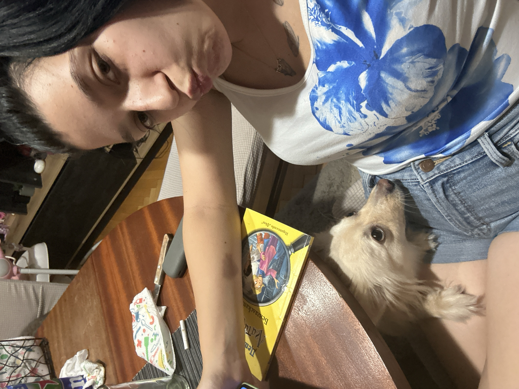 In a dimly lit indoor setting, a young woman with dark hair pulled back in a bun is taking a selfie. She has a playful expression, puckering her lips. She is wearing a white t-shirt with a large blue floral design and denim shorts. A small, light-colored dog with fluffy fur is nestled in her lap, looking at the camera. The woman's arm is resting on a yellow book with an illustration of a girl looking through a magnifying glass. A wooden table is visible in the foreground, with a glass and some napkins on it. The scene appears to be in a home environment, possibly a dining room.