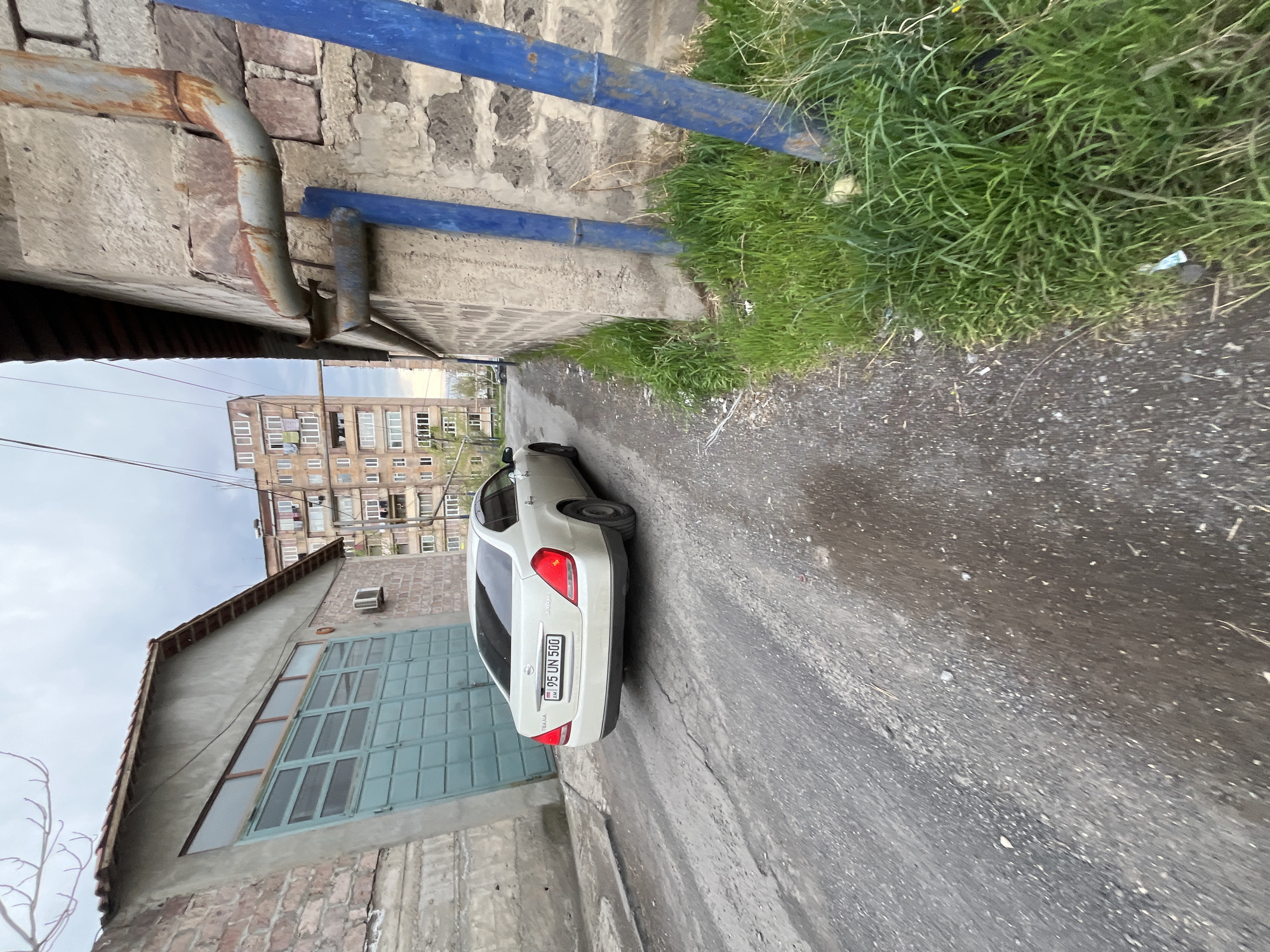 A street scene in Yerevan, Armenia, depicts a narrow alleyway with a parked white Nissan Teana bearing the license plate "95 UN 500". The alley is bordered by a brick and concrete building with a large, multi-paned green door on the left. On the right, a concrete wall with blue painted pipes and overgrowth of green grass is visible. The street surface is a gray asphalt texture. In the background, a multi-story residential building is visible under a cloudy sky. The image is taken at an angle, skewing the perspective.
