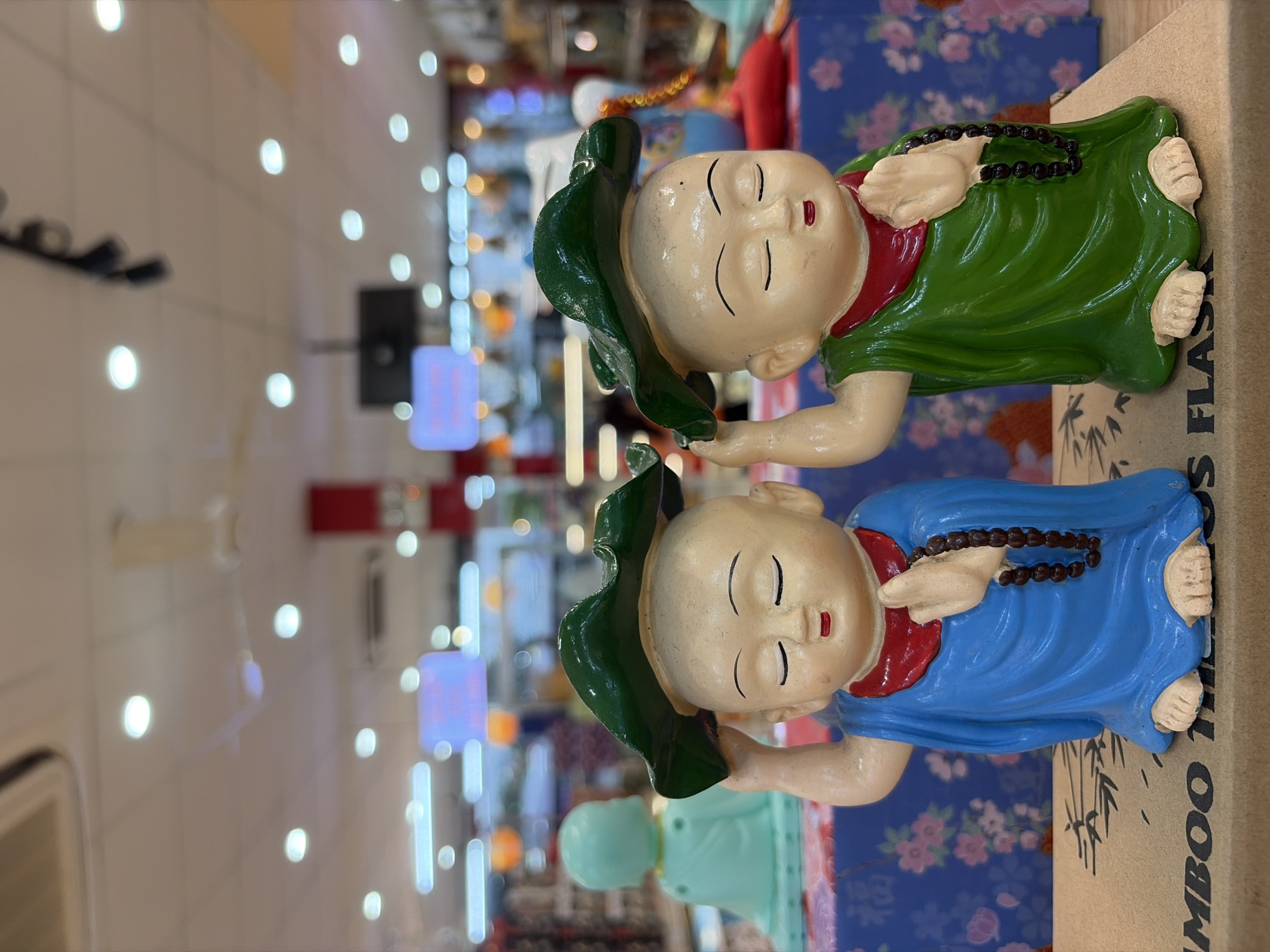 Two small statues of Buddhist monks are depicted from the waist up, with their eyes closed and serene expressions. Both are wearing green lotus leaf-like hats over their bald heads. The monk on the left is dressed in a blue robe with a red collar and a brown beaded necklace. The monk on the right wears a green robe with a red collar and a similar brown beaded necklace. They are standing side-by-side on a cardboard surface with Chinese characters visible, suggesting they are on display for sale. In the background, out-of-focus store lighting and merchandise can be seen, indicating an indoor retail environment. The specific location in Đà Nẵng, Vietnam cannot be determined from the image alone.
