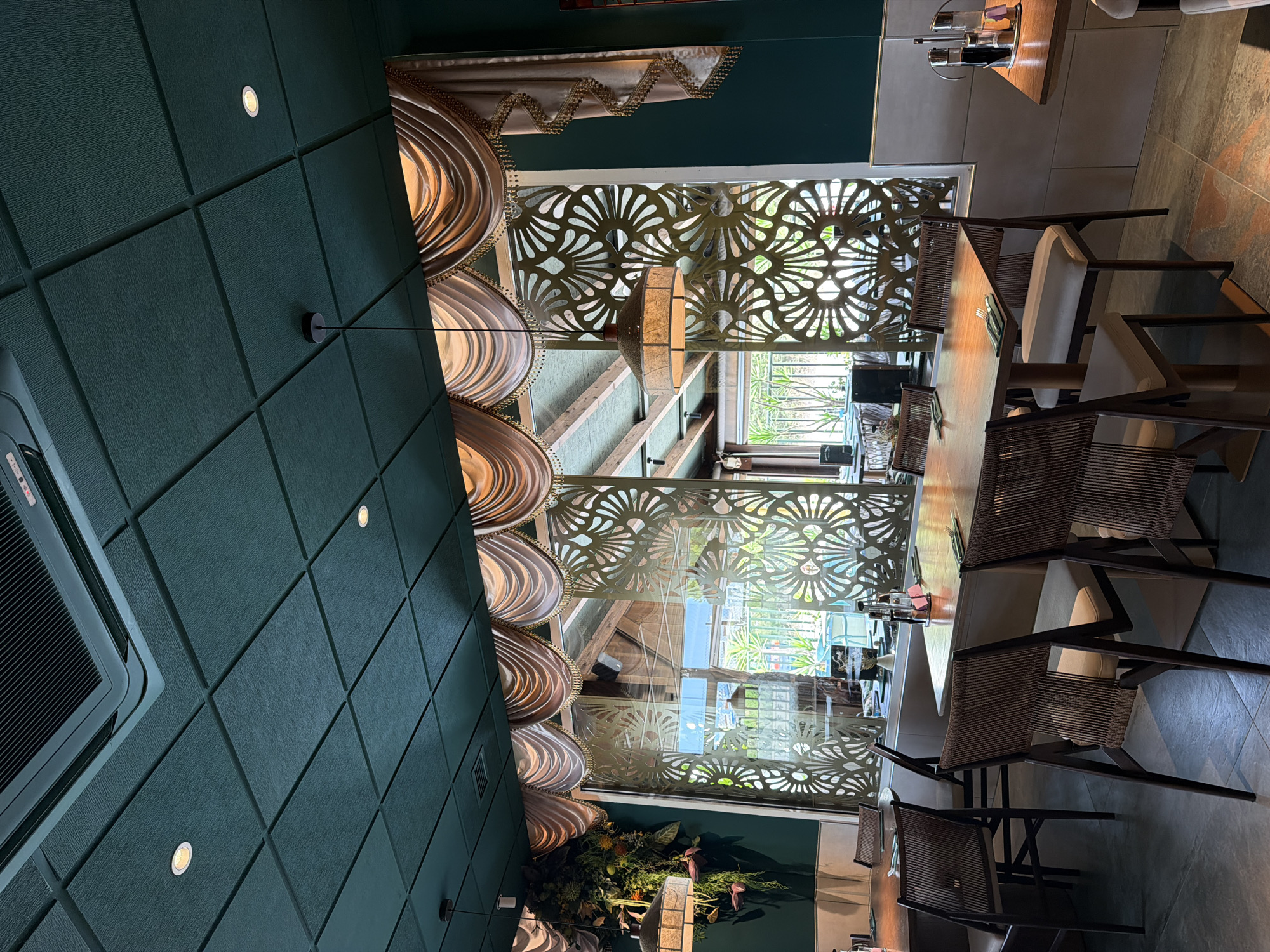 The image depicts an interior scene of a restaurant or dining area. The focal point is a long wooden table with chairs, suggesting a meal or gathering is either in progress or has recently concluded. The chairs appear to be made of dark wood with woven or rattan backs and light-colored upholstered seats. The table is set with cutlery and what appear to be condiment bottles.
The environment features a striking decorative screen with an intricate, geometric floral pattern, allowing light to filter through. This screen is situated behind the table, partially obscuring a view of the outdoors, which includes greenery and some architectural elements, possibly a balcony or another part of the building.
Above the table area, there are elaborate, decorative hanging elements that resemble draped fabric with golden trim and lighting, contributing to a luxurious ambiance. The ceiling is paneled with dark green textured material, interspersed with small circular lights. On the left side of the image, part of an air conditioning unit is visible.
The lighting in the scene is warm and diffused, suggesting it could be evening or an overcast day. The overall aesthetic is one of sophisticated dining with a touch of ornate design. There is no visible text in the image. The image is oriented vertically, with the scene appearing upside down relative to typical viewing.