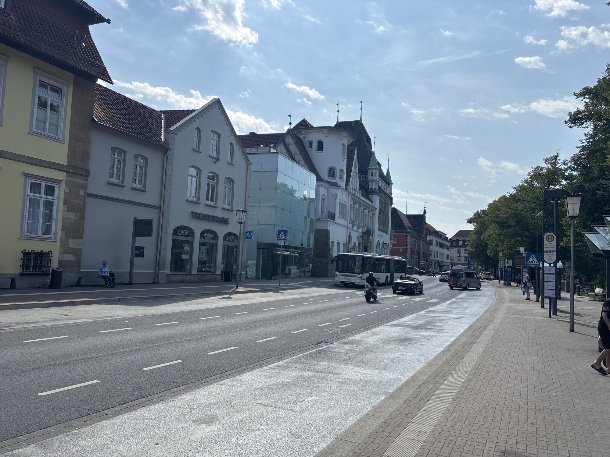 This image captures a street scene in Celle, Germany, likely on the street "Am Exer". On the left, a row of buildings lines the street, featuring a yellow building with a brown tiled roof and white-framed windows, followed by a light gray building with a similar roofline. The gray building has multiple windows, some with decorative ironwork, and a shopfront with the text "THEATERKASSE" visible. A person is seated on a bench in front of this building, wearing a blue shirt and dark pants.
The street itself is asphalt with white lane markings and a distinct white pedestrian crossing area. A motorcycle with a rider wearing a helmet is in the foreground, traveling in the same direction as a white bus further ahead. To the right of the street, a paved sidewalk is visible, bordered by trees and streetlights. Several bus stop signs are present, including one with the letter "H" indicating a bus stop. People are visible on the sidewalk, some waiting at the bus stop and others walking. A dark car is parked on the right side of the street. The sky is blue with scattered white clouds, and the sunlight casts shadows, indicating a daytime scene. The architectural style of the buildings suggests a historical European city center.