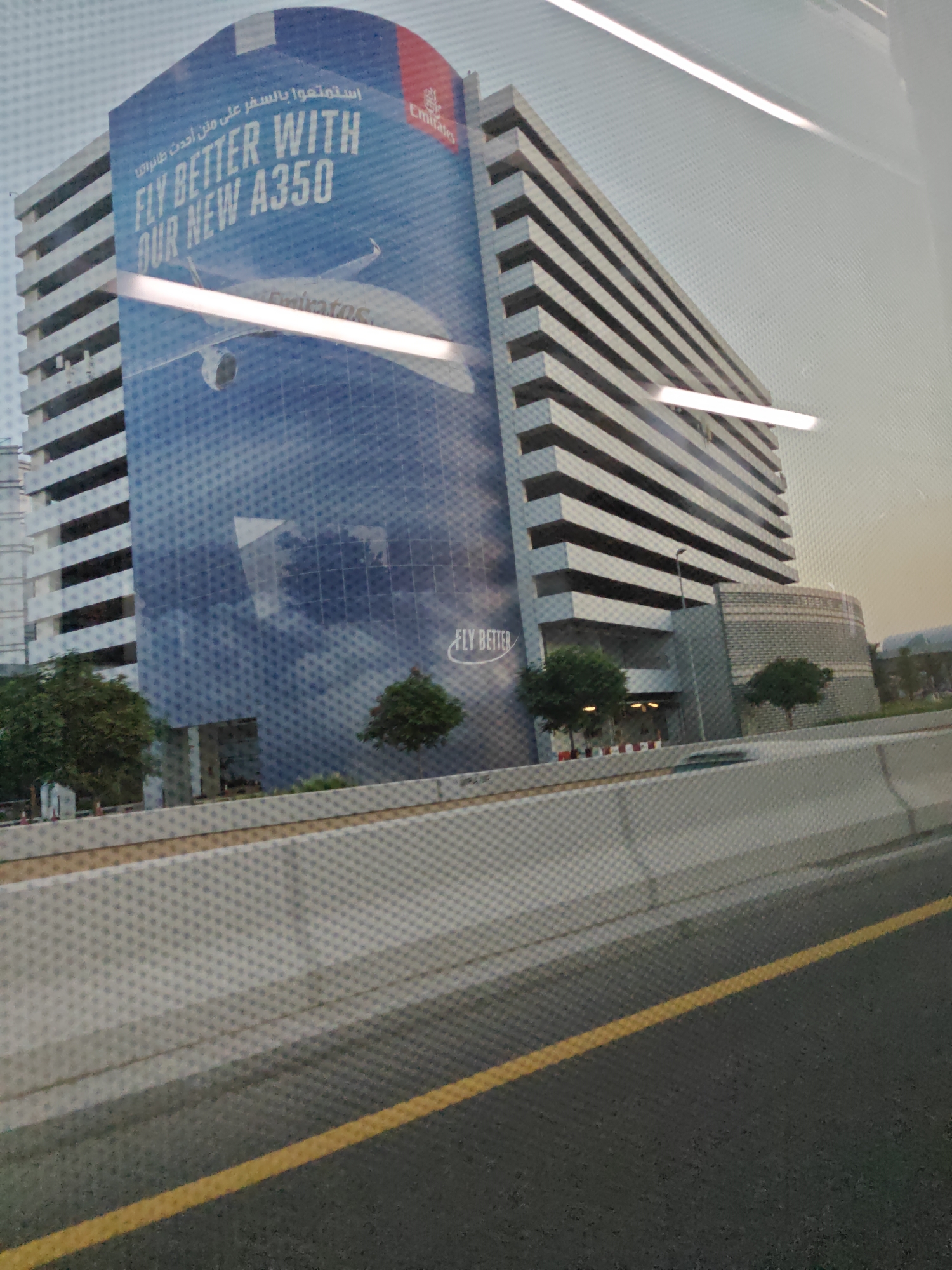 The image shows a multi-story building in Dubai, United Arab Emirates, that appears to be a parking garage. The side of the building is covered with a large advertisement for Emirates airline, featuring an image of an airplane and the text "FLY BETTER WITH OUR NEW A350". The advertisement also includes Arabic text and the Emirates logo. In front of the building is a highway with a yellow lane marker, and a concrete barrier separates the highway from the building. There are trees and some landscaping in front of the building. The overall scene depicts a typical urban environment in Dubai.