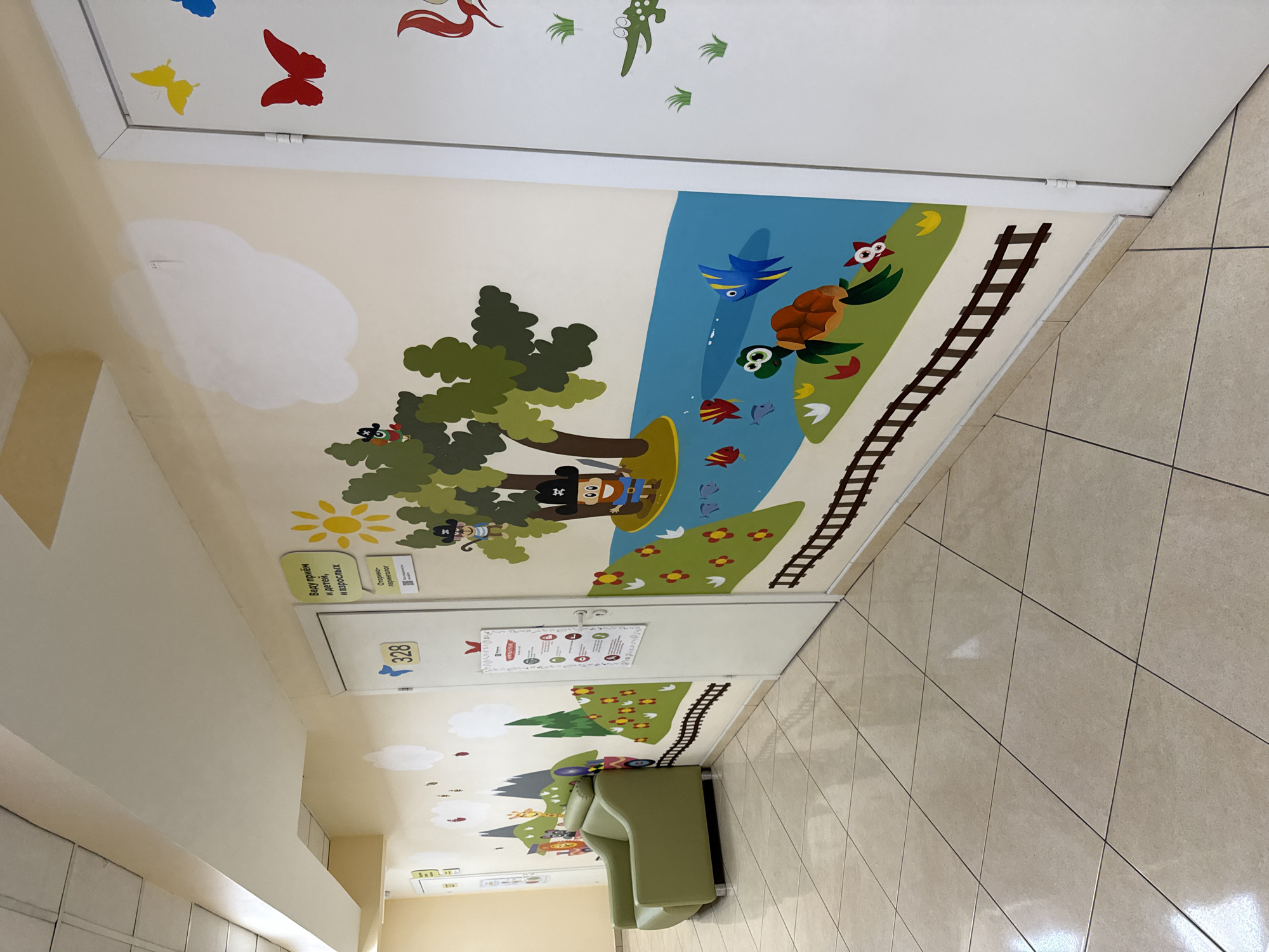 This image captures a brightly decorated corridor, likely within a children's medical facility or clinic in Moscow, Russia, given the cheerful murals and Russian text. The perspective is angled, looking down the hallway.
The walls are adorned with extensive, child-friendly cartoon murals depicting a vibrant natural landscape. On the upper wall, white clouds float across a pale yellow sky. Below, a green-leafed tree shades a river where a character resembling Mickey Mouse dressed as a pirate and a monkey in pirate attire float on a raft. The river section also features a blue bird, a turtle, a green frog, and several fish. Adjacent to the river, a grassy area is dotted with flowers and traversed by brown train tracks. The upper portions of the walls and ceiling also feature cutouts of butterflies and stylized green plant motifs.
Two white doors are visible along the corridor. One door displays the number "328" alongside a blue bird icon. Above this door, a yellow sun decal is affixed to the wall. Further down the corridor, another door has a poster with unreadable text and a sign above it that reads in Russian: "Ведет прием и детей, и взрослых" (Receives both children and adults) and "Открыто - перерыв!" (Open - break!). Below this text is a QR code and the small inscription "Docdoc.ru".
A light green upholstered bench or small sofa is positioned against the wall at the far end of the corridor, suggesting a waiting area. The floor is tiled with large, light-colored rectangular tiles laid in a diagonal pattern. The ceiling is plain white.
No people are present in the image, so no specific activity is taking place. The scene conveys a calm, inviting, and stimulating environment designed for young visitors. The lighting appears to be artificial and bright, illuminating the colorful decor.