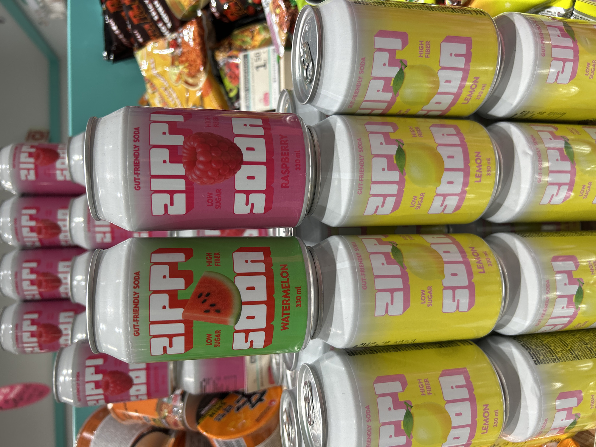 The image shows a close-up, eye-level, indoor shot of several cans of Zippi brand soda stacked on shelves in what appears to be a grocery store or convenience store. The cans are primarily in two flavors: watermelon and raspberry, distinguished by their can colors (green for watermelon, pink for raspberry) and corresponding fruit imagery. Several cans of a third flavor, lemon, are visible in the foreground and lower sections of the image, distinguished by their yellow cans and lemon imagery. The cans have white tops and silver pull tabs. In the background, other food products, including bags of chips and possibly more soda cans, are visible. The overall lighting suggests an indoor retail environment. The specific location in Matosinhos, Portugal, is not discernible from the visual information present in the image.