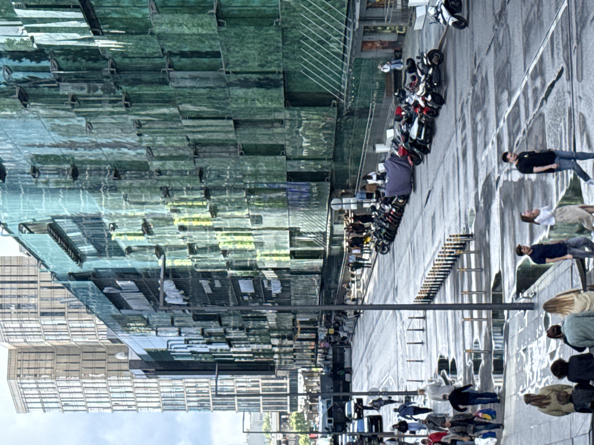The image depicts a street scene in Moscow, Russia, likely during daylight given the shadows. The main subjects are a group of people and a line of parked motorcycles. The setting is urban, with a modern glass-fronted building dominating the left side of the frame and a wet street on the right. The street appears to have been recently rained on, contributing to the reflective surfaces.
Several individuals are visible walking along the sidewalk or standing near the motorcycles. One man in a black t-shirt and jeans stands looking towards the viewer. Another person is seated on a motorcycle, and others are gathered in small groups. The motorcycles are a mix of styles and colors, predominantly dark.
The glass building reflects the sky and surrounding environment, creating a complex and somewhat distorted view of the scene. The overall impression is one of everyday urban life, with people going about their business in a modern cityscape. There are no visible signs with text.