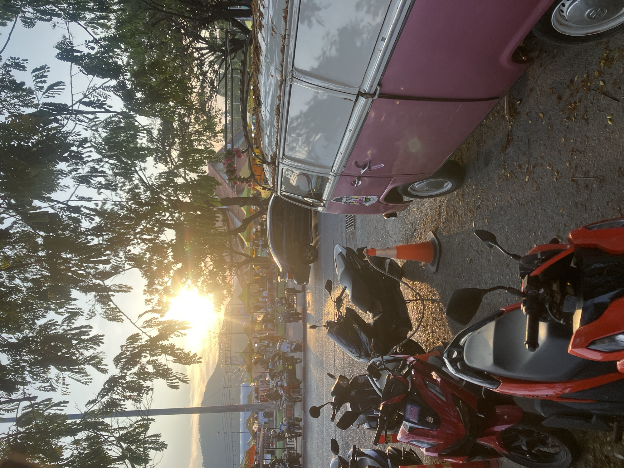 The image captures an outdoor scene at sunset or golden hour, characterized by warm, intense light filtering through tree branches. In the foreground, a vintage, two-tone pink and white Volkswagen Type 2 van (often called a Kombi or Bus) is parked on the left. Its roof is partially obscured by trees and appears to have some debris or decoration. To the right of the van, several motorbikes are parked; the closest one is red and black, showing a white sticker with the numbers "2570" on its side panel, while others are predominantly black. An orange traffic cone stands near the motorbikes.
Large trees with dense foliage frame the top and left side of the frame, silhouetted against the bright sky and the setting sun, which creates a prominent lens flare on the right. The ground is paved asphalt, strewn with fallen leaves. In the mid-ground and background, under the warm glow of the sun, a bustling market or fairground is visible, featuring numerous stalls, tents, and a distant Ferris wheel. Mountains rise faintly in the far background beneath a soft sky. The scene suggests a lively, festive atmosphere in Baan Chaweng, Thailand, as the day transitions to evening.