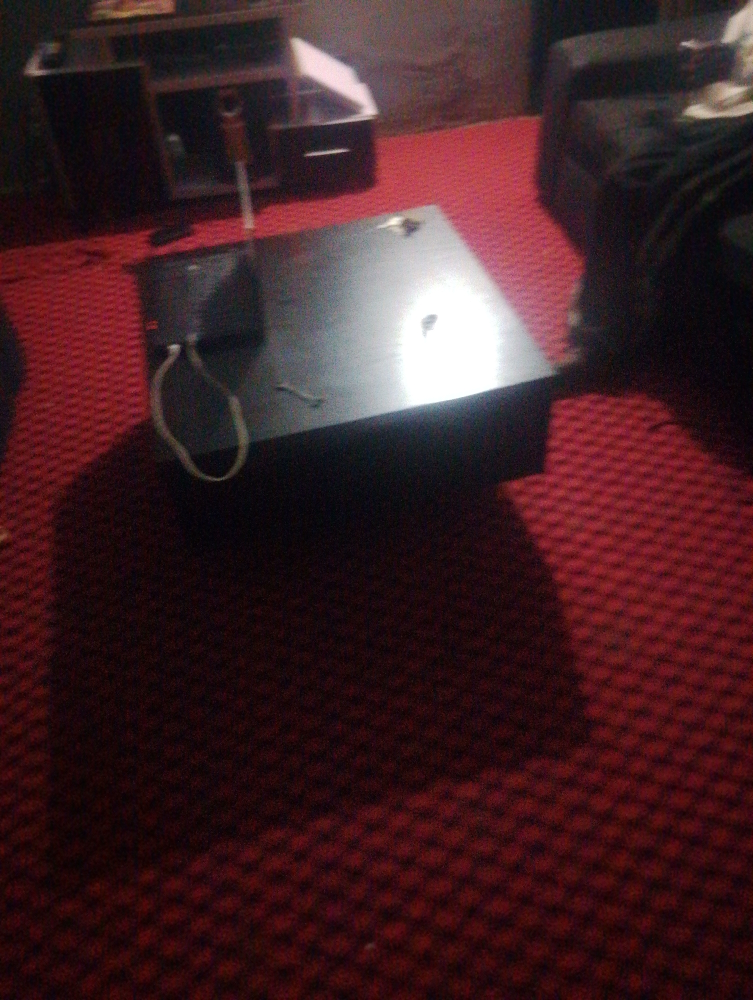 The image shows a low-angle, dimly lit view of a low-profile black coffee table situated on a textured red carpet. A thin, light-colored cord or strap is draped over the left edge of the coffee table, forming a loop. The background is dark and out of focus, suggesting an indoor setting, possibly a living room or lounge area. There is no indication of the specific location within the city of Jos, Nigeria, as the image lacks any discernible landmarks or contextual clues. No people or actions are visible.
