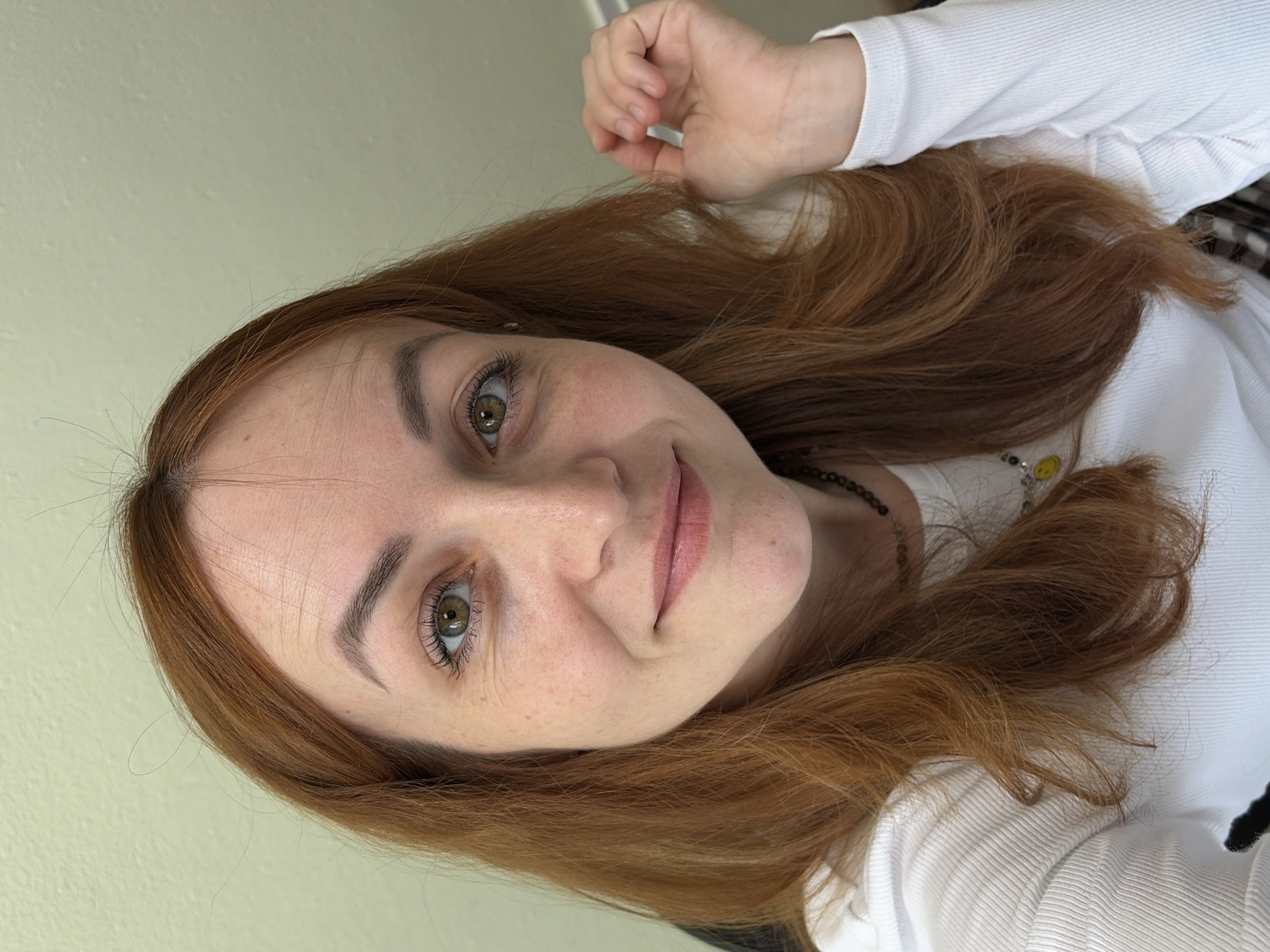 A close-up, rotated portrait shows a young woman with long, wavy reddish-brown hair and light green/hazel eyes, looking directly at the viewer with a gentle smile. Her fair skin has subtle freckles across her nose and cheeks. She is wearing a white, ribbed long-sleeved top. A dark necklace with a small, round yellow charm featuring a black smiley face is visible around her neck, partially covered by her hair. Her left hand, with a closed fist, is positioned near the top of her head. The background is a plain, light, neutral-colored wall, suggesting an indoor setting. The image, taken in Chaikovsky, Russia, captures a moment of calm and approachability.