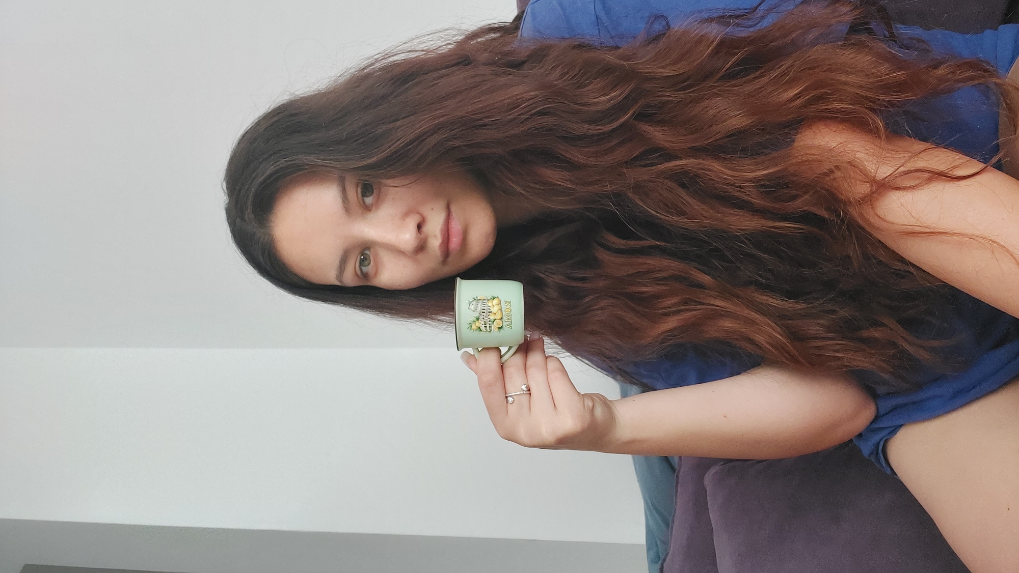 The image shows a young woman in an indoor setting, likely in Shumen, Bulgaria, during daytime. She is depicted in a reclining or lounging position, with her head slightly tilted and her gaze directed calmly towards the camera. Her long, wavy hair, a rich reddish-brown color, spreads around her. She is dressed in a casual blue short-sleeved top.
In her right hand, she holds a small, light green ceramic cup. The cup is adorned with a yellow lemon design and the word "ROMY" printed vertically on its side. On her right ring finger, she wears an open-ended ring featuring two clear, sparkling stones.
The woman appears to be seated or leaning against soft furniture, possibly a couch or cushioned chair, which is upholstered in a deep purple or dark grey fabric and visible in the lower portion of the frame. The background consists of a plain, light-colored wall, likely white or light grey, indicating a simple home interior. The lighting is bright and even, suggesting natural daylight or good indoor illumination. The overall impression is one of relaxed repose.