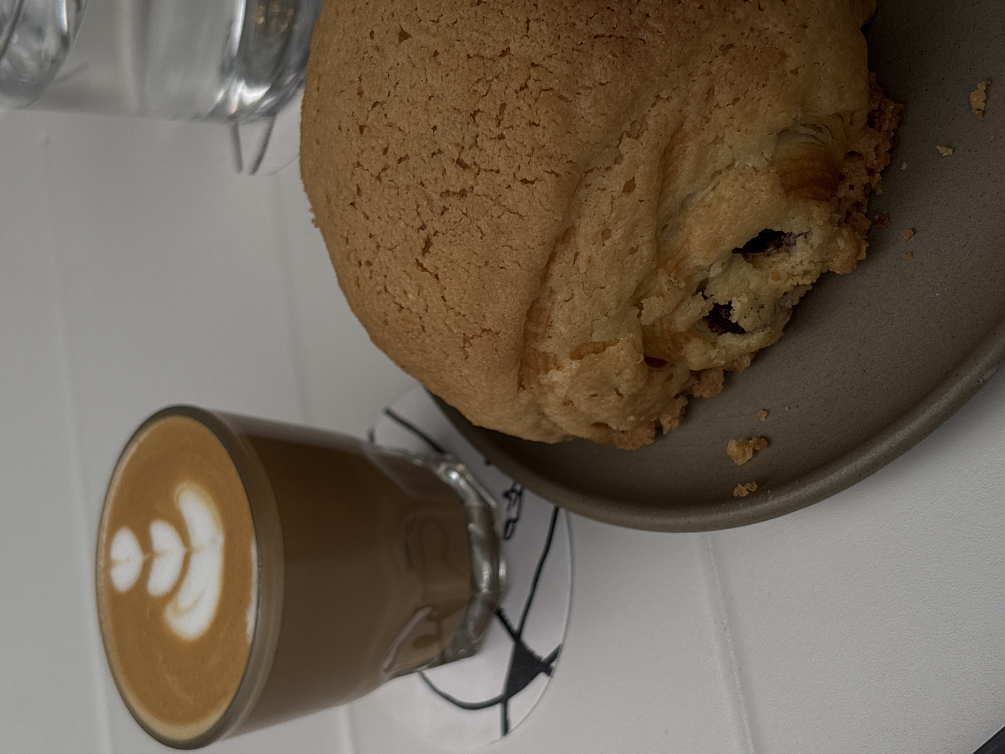 A top-down, slightly tilted shot captures a casual cafe setting in Mexico City, featuring a coffee and a large bun on a white table.
The main subject is a substantial, golden-brown bun, possibly a melon pan or similar sweet bread, positioned on a matte gray, round plate towards the upper right of the frame. Its surface is textured and appears crusty. A visible crack on one side reveals a dark filling, suggestive of berries or another fruit, within the softer interior of the bun. Scattered crumbs are visible on the plate.
To the lower left, a clear glass contains a latte with intricate white latte art. The design features multiple layered heart or leaf shapes, indicating careful preparation. The glass rests on a white coaster adorned with a distinct black geometric pattern. In the upper left background, another glass object is partially visible and out of focus.
The scene suggests a moment of enjoyment, perhaps a breakfast or afternoon snack. The bright, even lighting indicates it is daytime. There is no visible text in the image.