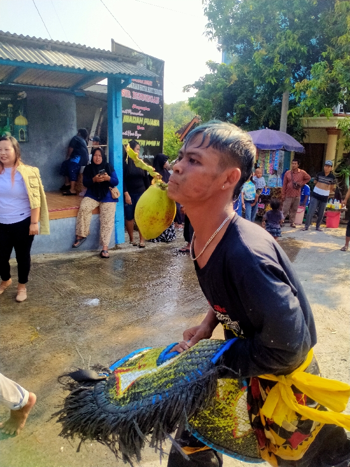 The image depicts an outdoor street scene in Sepat, Indonesia, under bright daylight. In the foreground, a male performer with blue-tinted hair, wearing a black long-sleeved shirt, a silver chain, and a yellow waist sash, holds a large, fringed, multi-colored ceremonial prop with both hands. He is simultaneously holding a green coconut by its husk in his mouth, his head tilted back slightly. The paved ground beneath him is wet. To the left, a woman in a white shirt and khaki jacket observes the scene, while further back, another woman in a blue shirt and patterned pants sits on a raised concrete platform, looking at a mobile phone. This platform is part of a building with blue support columns and a corrugated metal roof, featuring a sign with Indonesian text, including "IBADAH PUASA," and a large green mosque decal. To the right of the performer, a street vendor stall with a purple umbrella displays various packaged items, with several adults and children gathered around it. Trees and a distant blue building are visible in the background.