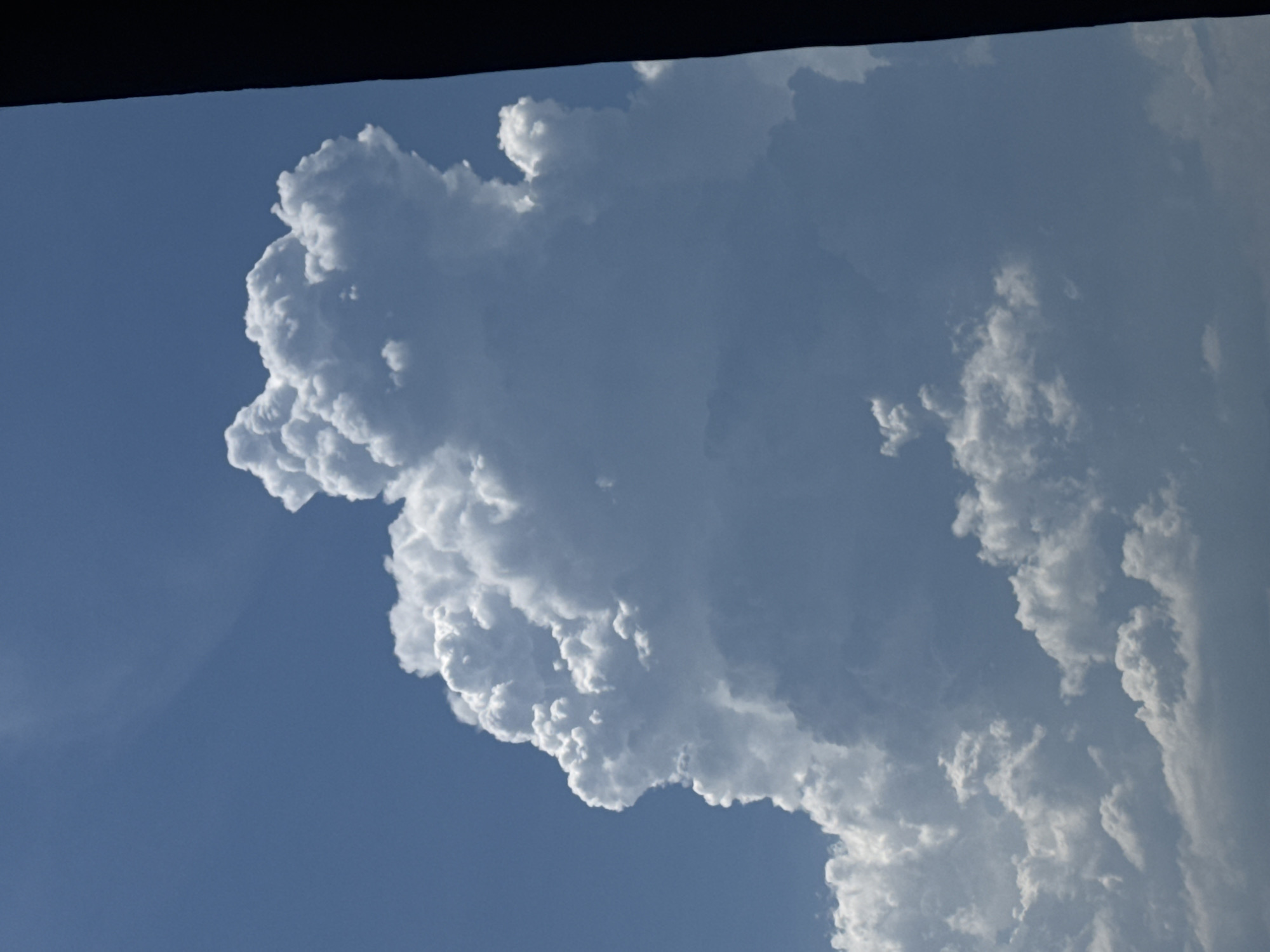 The image displays a dynamic sky with a large, dramatic cumulus cloud formation dominating the center. The sky is a clear, bright blue, suggesting a sunny day. The cloud is massive and textured, with bright white highlights on its upper portions where sunlight is directly hitting it, and softer, shadowed areas in its depths. The cloud appears to be a towering cumulonimbus cloud, potentially indicative of developing weather.
The lower portion of the image shows smaller, more scattered cumulus clouds at a lower altitude, appearing somewhat wispy and less defined than the main cloud.
On the right side of the image, a dark, vertical rectangular shape intrudes into the frame, obscuring a portion of the sky. This object appears to be a solid, dark structure, possibly a building or a frame element, seen from a low angle.
The image is an outdoor shot, likely taken during the daytime. There are no people or identifiable objects within the cloud formations or the blue sky itself. The scene is solely focused on the atmospheric conditions. The lighting suggests the sun is high in the sky, casting strong illumination on the clouds. The perspective appears to be looking upwards.
While the prompt asks for a location in Nha Trang Ward, Vietnam, there are no specific visual cues in the image to confirm this location. The scene is a generic depiction of a cloudy sky.