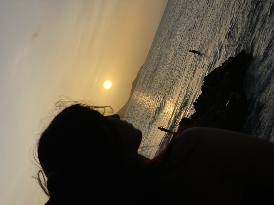 Silhouetted head and shoulder dominate the foreground, contrasting with the bright sunset over the ocean. The sun's reflection creates a shimmering path on the water. A rocky outcrop stands to the right. Two figures are visible in the distance; one standing near the rocks, the other further out in the water. The sky is pale orange, blending into the darker ocean. The image is rotated slightly counter-clockwise.