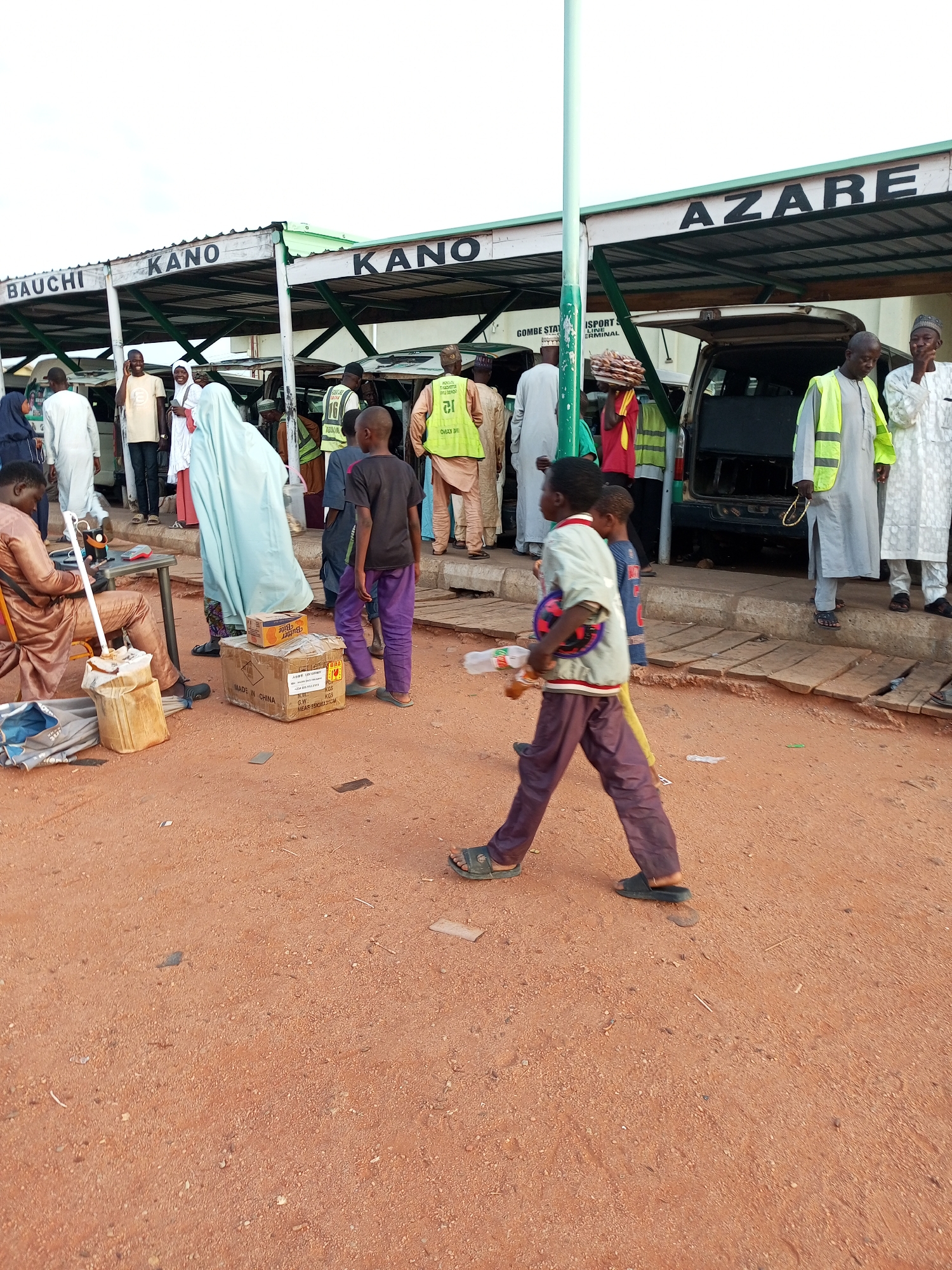 The image depicts an outdoor scene at a bus terminal in Gombe, Nigeria, under a cloudy sky. Several people are visible, some standing and some walking, near a covered waiting area. Signs above the covered area indicate destinations such as "BAUCHI," "KANO," and "AZARE." Vehicles, including vans, are parked behind the waiting area. In the foreground, a young boy in a light-colored shirt and dark pants walks from right to left, carrying a toy. Another child is walking behind him. To the left, a man in a brown robe is seated on a low stool next to some bags. Two cardboard boxes are on the ground in the middle of the scene. The ground is covered in reddish-brown dirt and scattered debris.