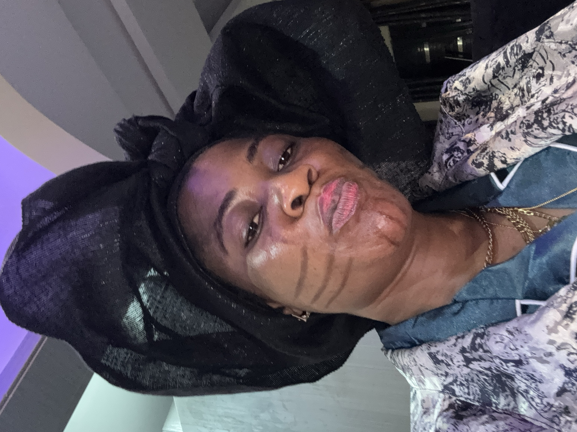 A close-up, selfie-style photograph features a dark-skinned woman from Ajara, Nigeria, wearing a large, elaborate black headwrap and a patterned jacket. The woman has distinct, dark lines drawn on her cheeks, resembling tribal markings, and her lips are pursed. She is wearing multiple gold necklaces. The background is dimly lit and appears to be an indoor setting with a textured wall and a hint of purple lighting from above.