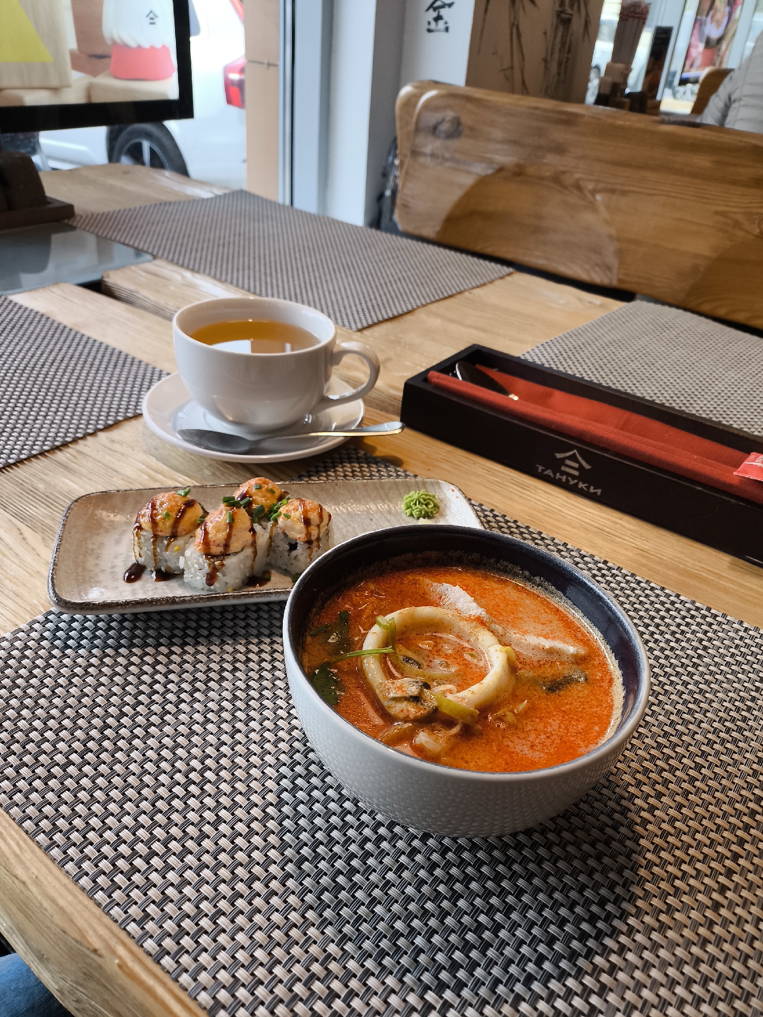 The image shows a meal at a restaurant or cafe, likely in Moscow, Russia based on the presence of "Tanuki" branded chopsticks, a popular sushi chain there. A table setting includes a bowl of red-colored soup (possibly Tom Yum), a small plate of sushi rolls topped with a creamy sauce and a drizzle, a dab of wasabi, a cup of tea, and a set of chopsticks in a black and red case. The table has a woven placemat and a light-colored wooden surface with inlays of darker wood. The background shows a window looking onto a street scene, including a car. The lighting suggests it is daytime. The scene is set indoors, and the focus is on the food arrangement on the table.