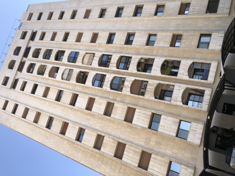 The image displays a multi-story residential building located in Kafr Sousa Municipality, Syria, viewed from a low, upward angle against a clear blue sky. The building features a light-colored, possibly limestone or tiled, facade with pronounced vertical columns separating recessed sections containing windows and balconies.
The structure appears to have at least 12 visible stories. Each story features a repeating pattern of rectangular windows and arched balconies. Many windows are fitted with light brown external roller shutters, while others are glass, reflecting the blue sky. The balconies, distinguished by dark metal railings, frequently house external air conditioning units. On one mid-level balcony, a green potted plant or small tree is visible, and on another lower balcony, light-colored laundry or textiles are hanging.
At the base of the building, a dark, arched carport or shade structure extends, underneath which a light-colored vehicle, possibly silver or light blue, is partially visible. The overall environment suggests a well-maintained urban residential area under bright daylight. No individuals are present in the scene.