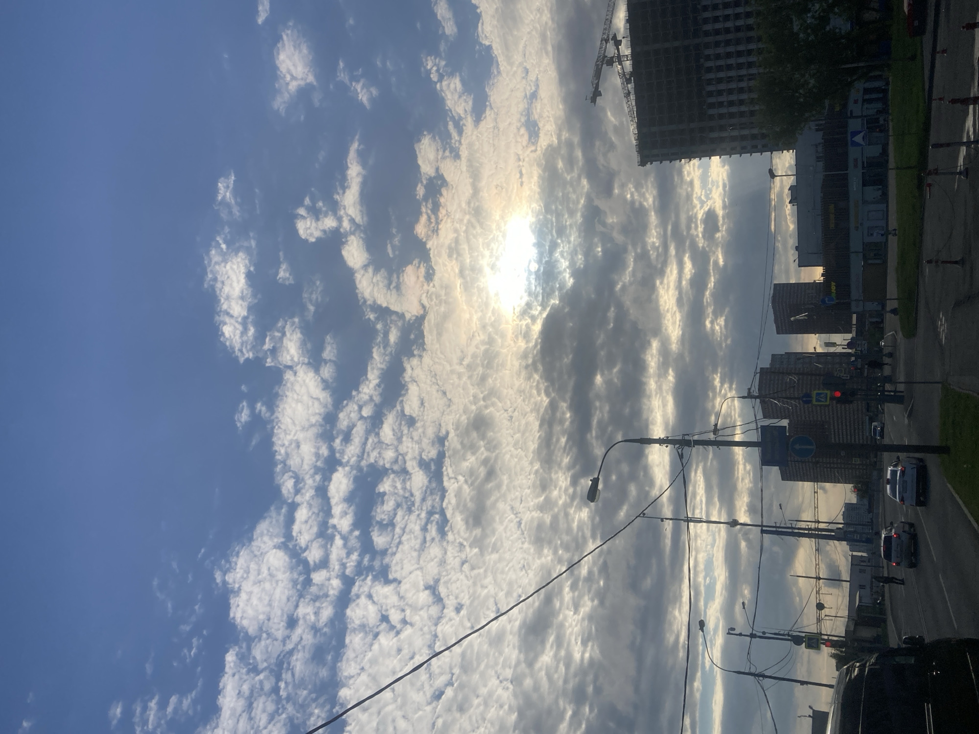 The image shows a view of a street scene in Moscow, Russia, under a partly cloudy sky with the sun shining through the clouds. A building under construction with a crane is visible. Several other buildings are visible along the street. There are streetlights and power lines. Cars are traveling on the road.