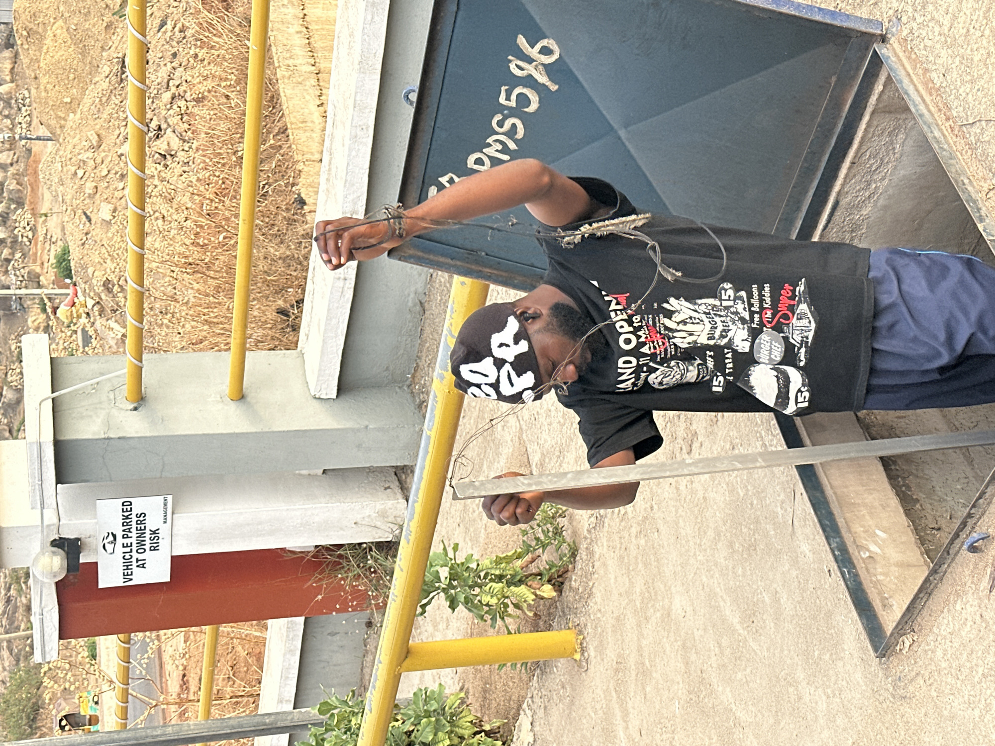 The image shows a man working on a utility access point, likely a manhole, in what is possibly Jos, Nigeria, based on the writing "PMS596" on the access cover which indicates the Petroleum Products Pricing Regulatory Agency (PPPRA) depot location in Jos. The man is wearing a black t-shirt with graphics, a patterned bandana on his head, glasses, and blue pants. He is holding a metal rod with wires attached to his upper arm. The utility access point is in a concrete surface and surrounded by yellow guard rails. A sign reading "VEHICLE PARKED AT OWNERS RISK MANAGEMENT" is attached to the railing structure. The surrounding environment appears to be a hilly, dry landscape with sparse vegetation.