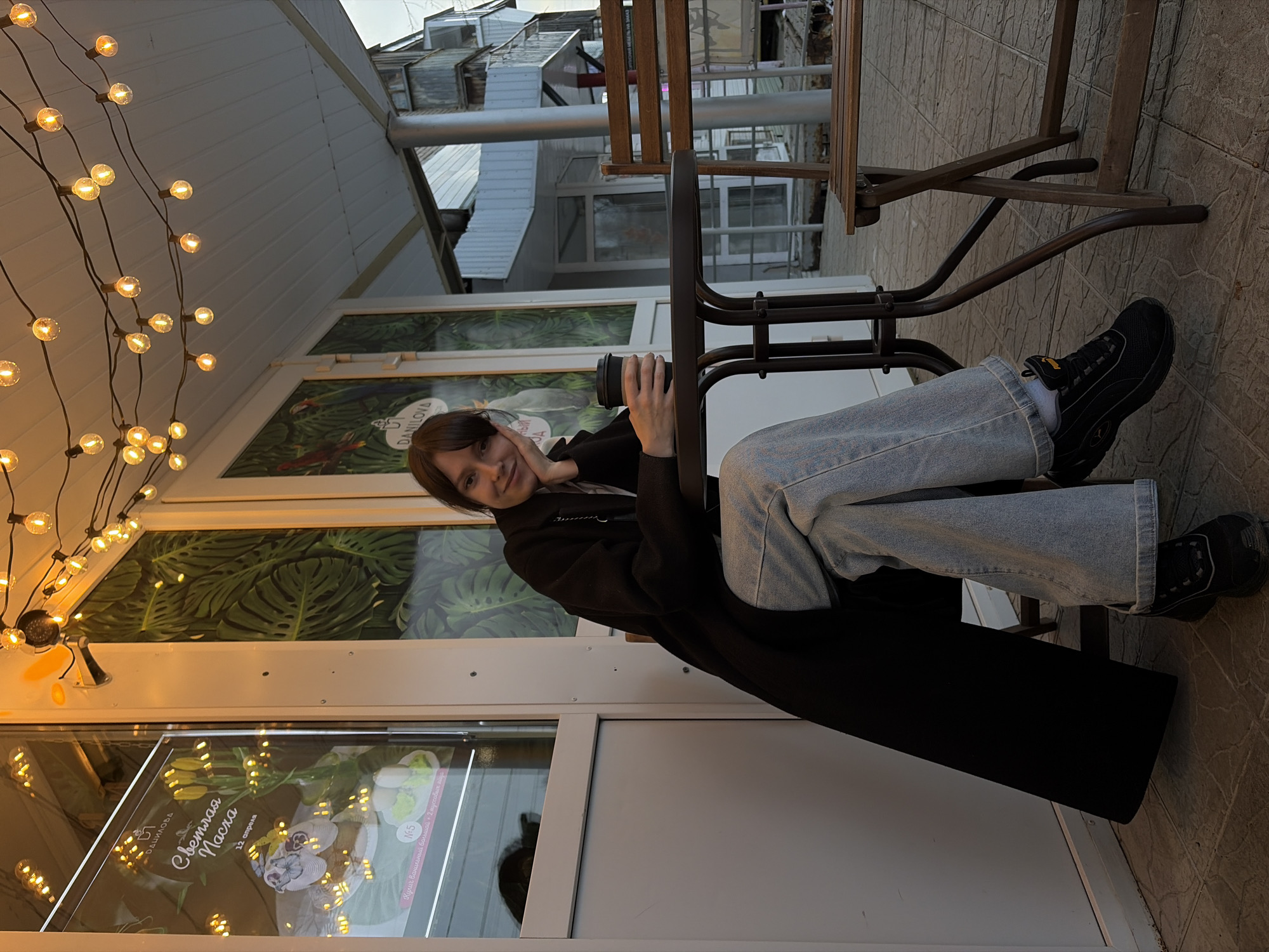 The image shows a young woman sitting at an outdoor table outside a cafe or shop. She is wearing light blue jeans, a dark top, and a long black coat. She is holding a dark-colored cup and resting her head on her hand, looking toward the camera with a slight smile.
The location appears to be Chaikovsky, Russia, based on the text "Чайковский" visible in the shop window ("Светлая Пасха. Чайковский"). The shop window displays posters and floral arrangements.
Behind the woman are windows decorated with a tropical leaf pattern. Strings of lights hang above, illuminating the scene. The building appears to be a commercial establishment. The flooring is tiled. Other tables and chairs are visible nearby. The sky is overcast.