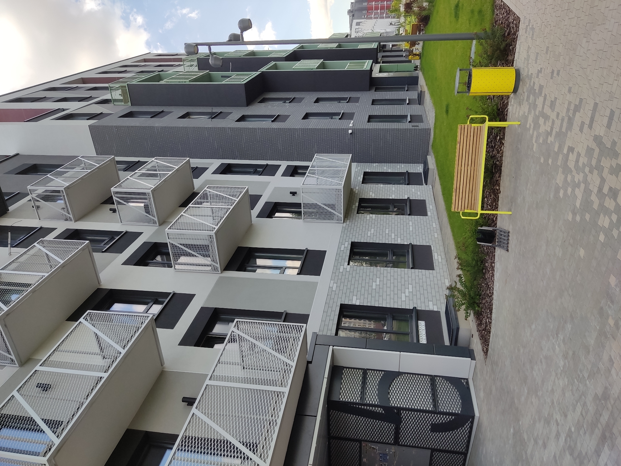 This is a medium-angle, exterior, eye-level shot of a modern apartment building in the city of Baravliany, Belarus. The building is characterized by its dark grey and light grey facades, interspersed with white balconies that feature intricate geometric metal railings. Several floors of the building are visible, with numerous dark-framed windows. The ground level features a dark metal entrance gate with a patterned design. In front of the building, there is a paved walkway made of interlocking grey pavers, leading to a grassy area. A bright yellow bench with wooden slats and a matching yellow cylindrical trash bin are positioned on the grass. A street lamp with multiple heads stands to the right of the bench. The sky is partly cloudy with a mix of blue and white. The scene appears to be devoid of people.