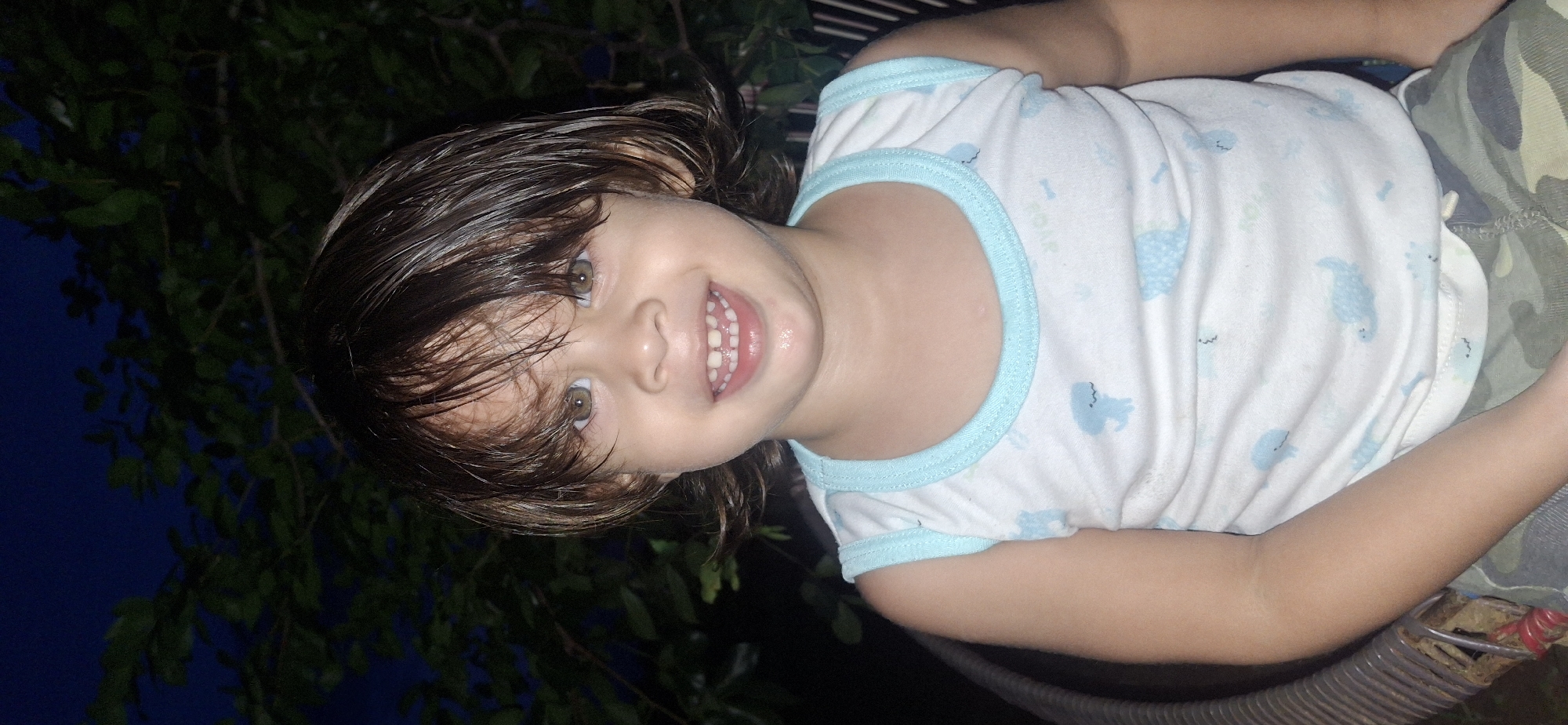 A close-up, waist-up shot shows a young child with light brown hair and hazel eyes, smiling at the camera. The child is wearing a white tank top with light blue trim and a pattern of small blue dinosaurs. The background is dark and appears to be outdoors at night, with dark green leaves and branches visible against a deep blue sky. The child is seated in what appears to be a chair with a woven texture. The scene is likely in Vallecito, Venezuela, given the context.