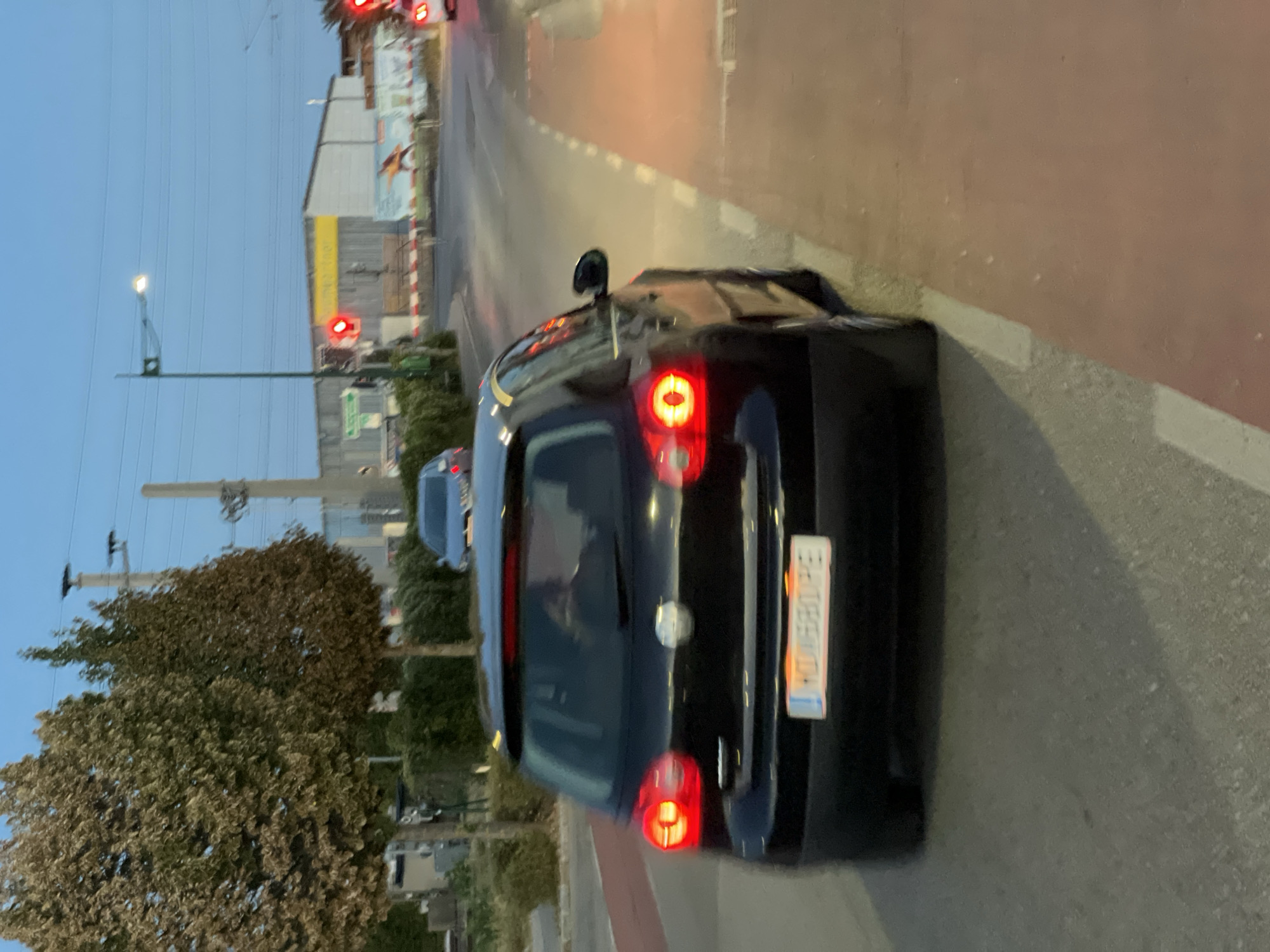 The image shows a black Volkswagen Golf driving on a street in Mödling, Austria. The car's license plate reads "MD680PE". It is daytime, and the sky is a clear blue. There are trees and buildings in the background, and traffic lights are visible. The street appears to be a regular city street with a bike lane marked in red. The car is in motion, and the image is slightly blurred, indicating it was taken from another moving vehicle.