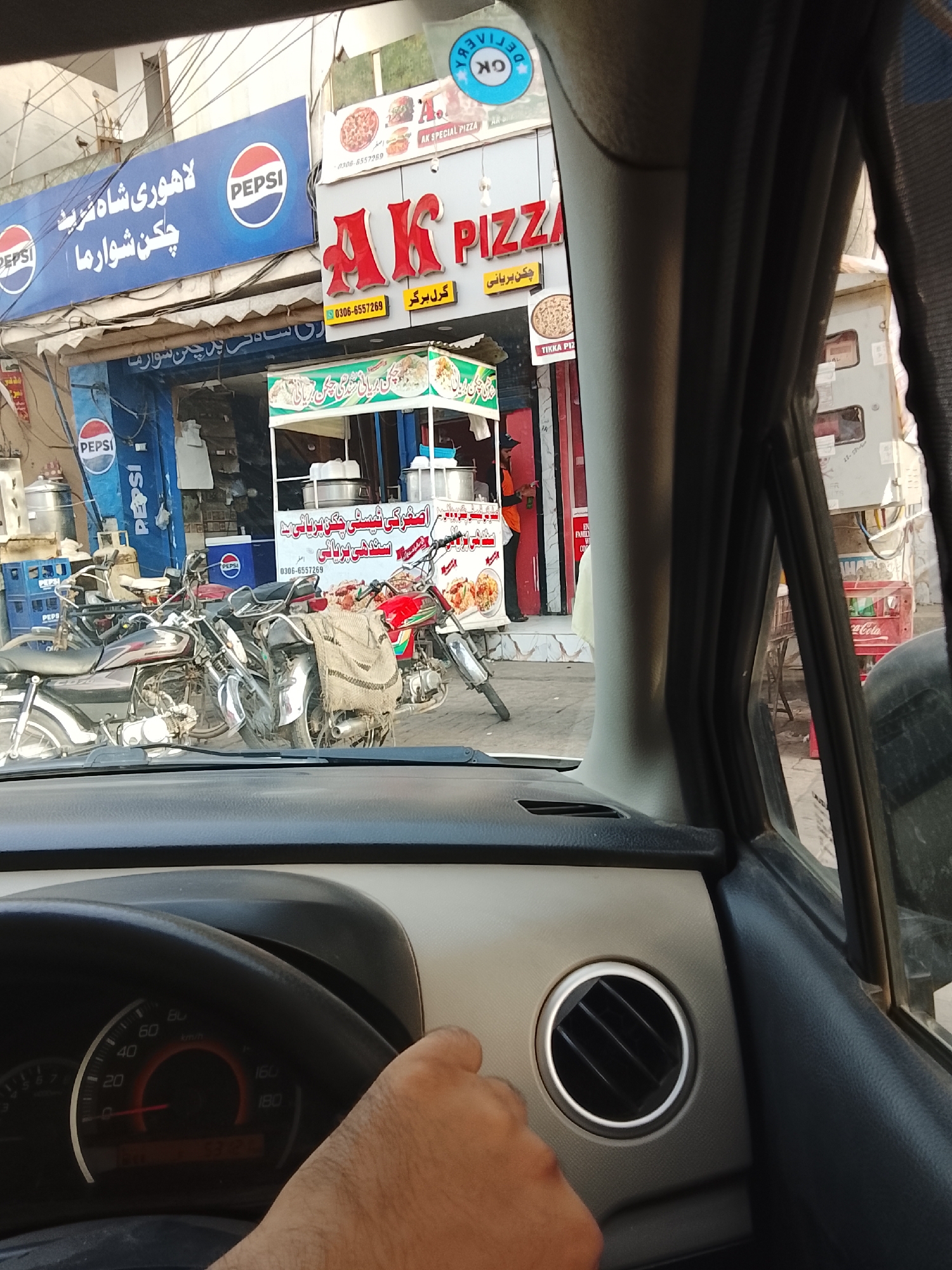 The image, taken from inside a car, shows a street scene in Gulgasht Colony, Pakistan. A sign for "Lahori Shah Sharif Chicken Shawarma" is prominently displayed above a Pepsi advertisement. Next to it, there's a sign for "AK Pizza" and "AK Special Pizza." A food cart selling items like "Asghar's Chicken Biryani" is visible in front of the shops. Several motorcycles are parked on the sidewalk. The car's dashboard and steering wheel are in the foreground, with the driver's hand visible. The environment appears urban, with commercial establishments dominating the street.