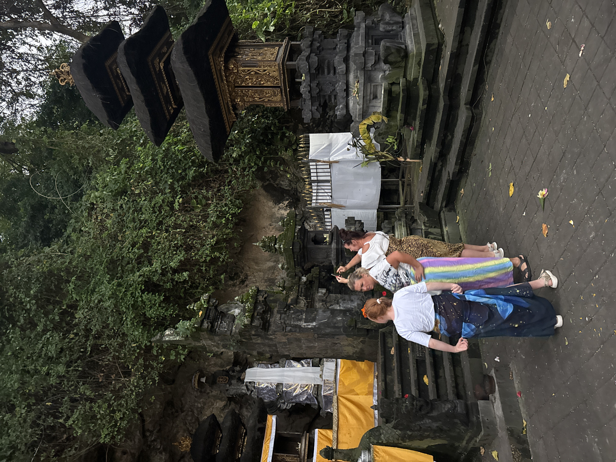 In Dawan, Indonesia, three women are gathered in front of a stone temple structure. The temple features multiple tiers topped with black thatched roofs and ornate golden decorations. To the left, another temple structure is visible with yellow fabric draped around its base. The ground is paved with dark stone tiles, and fallen leaves are scattered across it. Lush green foliage covers the rocky hillside behind the temples. One woman, on the right, is taking a photo with her phone. Another woman in the center is looking towards the temple, while the woman on the left is adjusting her sarong. The lighting suggests it is late afternoon or early evening.