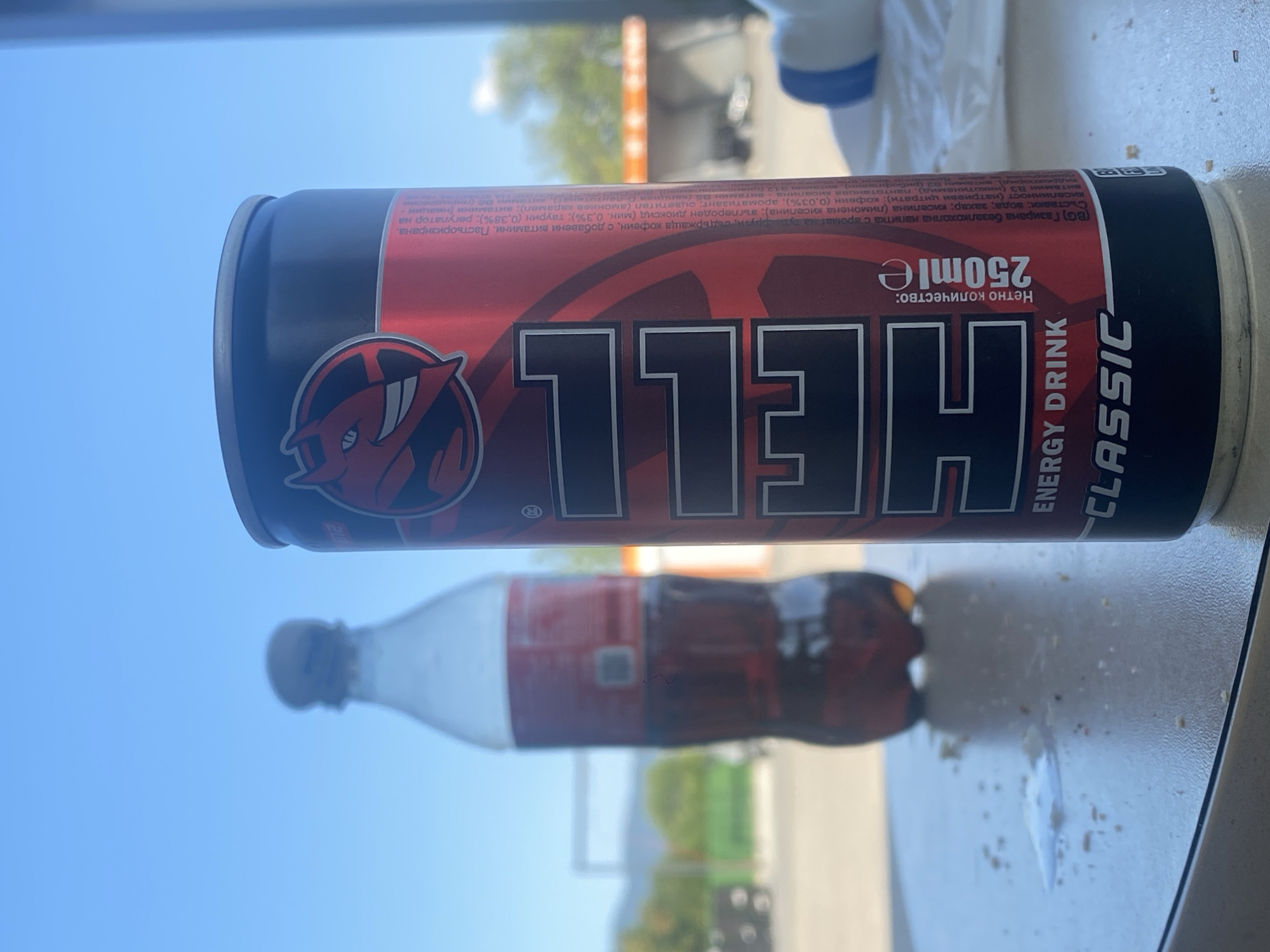 An outdoor scene with a blue sky dominates the background. In the foreground, a red and black can of "HELL ENERGY DRINK CLASSIC" stands upright on a speckled white surface, possibly a table. The can is partially obscured by its own label, which prominently features the "HELL" logo with a red devil icon. To the left and slightly out of focus is a plastic bottle filled with a dark brown liquid, likely a beverage. The surface also shows scattered crumbs and a small puddle, suggesting a recent consumption or spillage. The background is blurred but hints at an urban or semi-urban environment with some structures and greenery. The setting appears to be the city of Kran, Bulgaria.