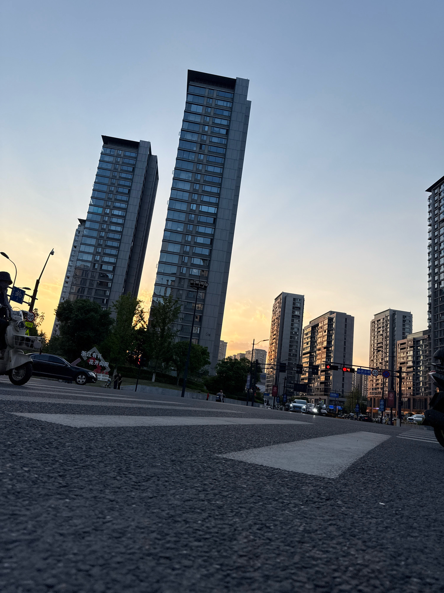 This is a low-angle, wide-shot photograph of a city street scene in Yiwu, China, during what appears to be either sunrise or sunset. The foreground is dominated by asphalt road with prominent white pedestrian crossing lines. In the midground, several modern, tall residential buildings with numerous windows line the street. Some of these buildings are leaning slightly due to the camera's perspective. A few trees are visible between the buildings and the road.
On the left side of the frame, a person is riding a white scooter. Traffic lights and street lamps are present at an intersection. Several cars and a few pedestrians are visible on the road and sidewalks. A banner with Chinese text is displayed near one of the buildings. The sky in the background is a gradient of light blue to orange, indicating the low angle of the sun.