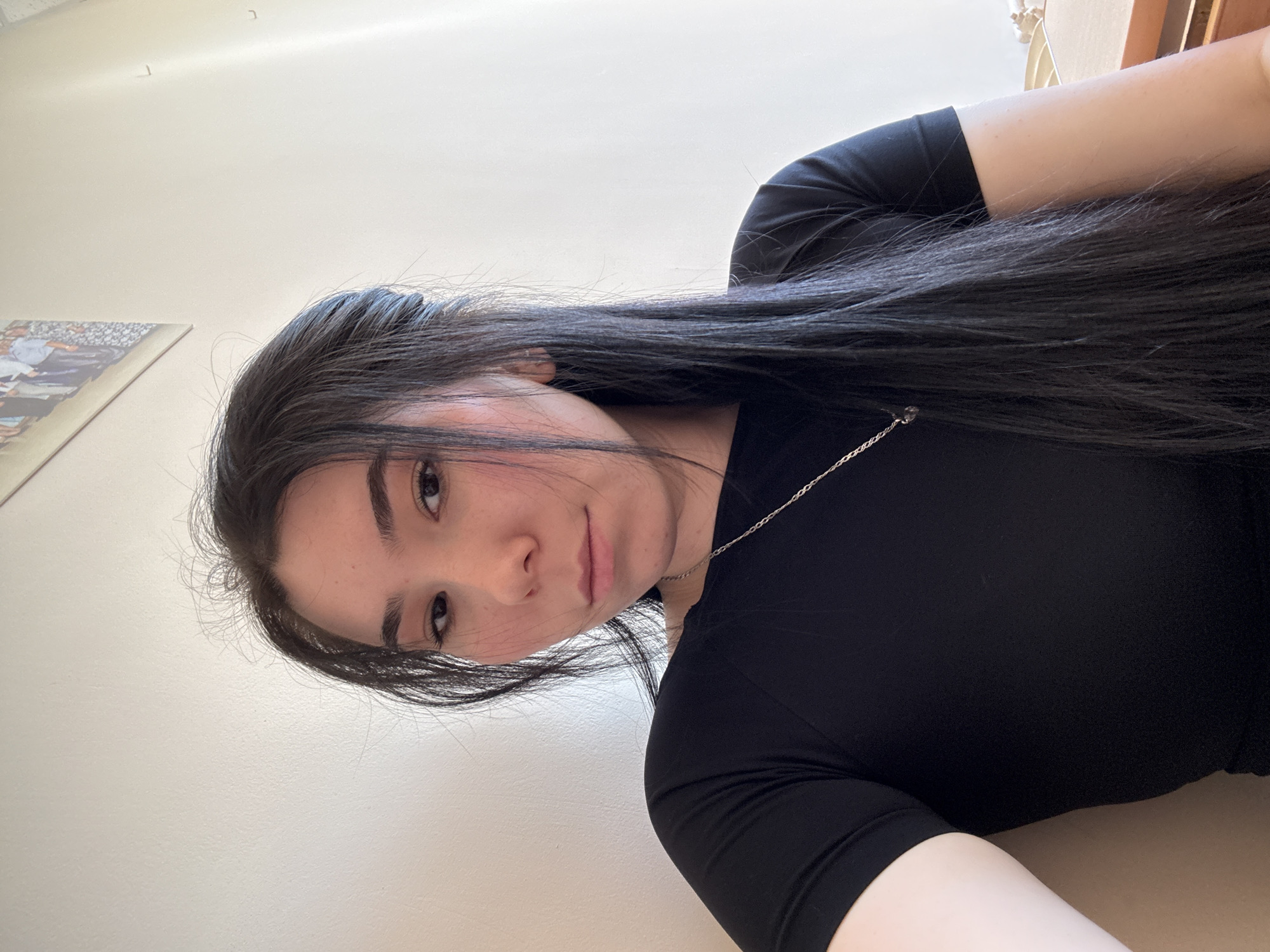 A medium shot of a fair-skinned woman with long, straight black hair, wearing a black top and a silver chain necklace. She is facing the camera with a slight tilt of her head. A framed picture hangs on the white wall behind her. The lighting appears natural. There is no contextual information to definitively place the scene in Izhevsk, Russia.