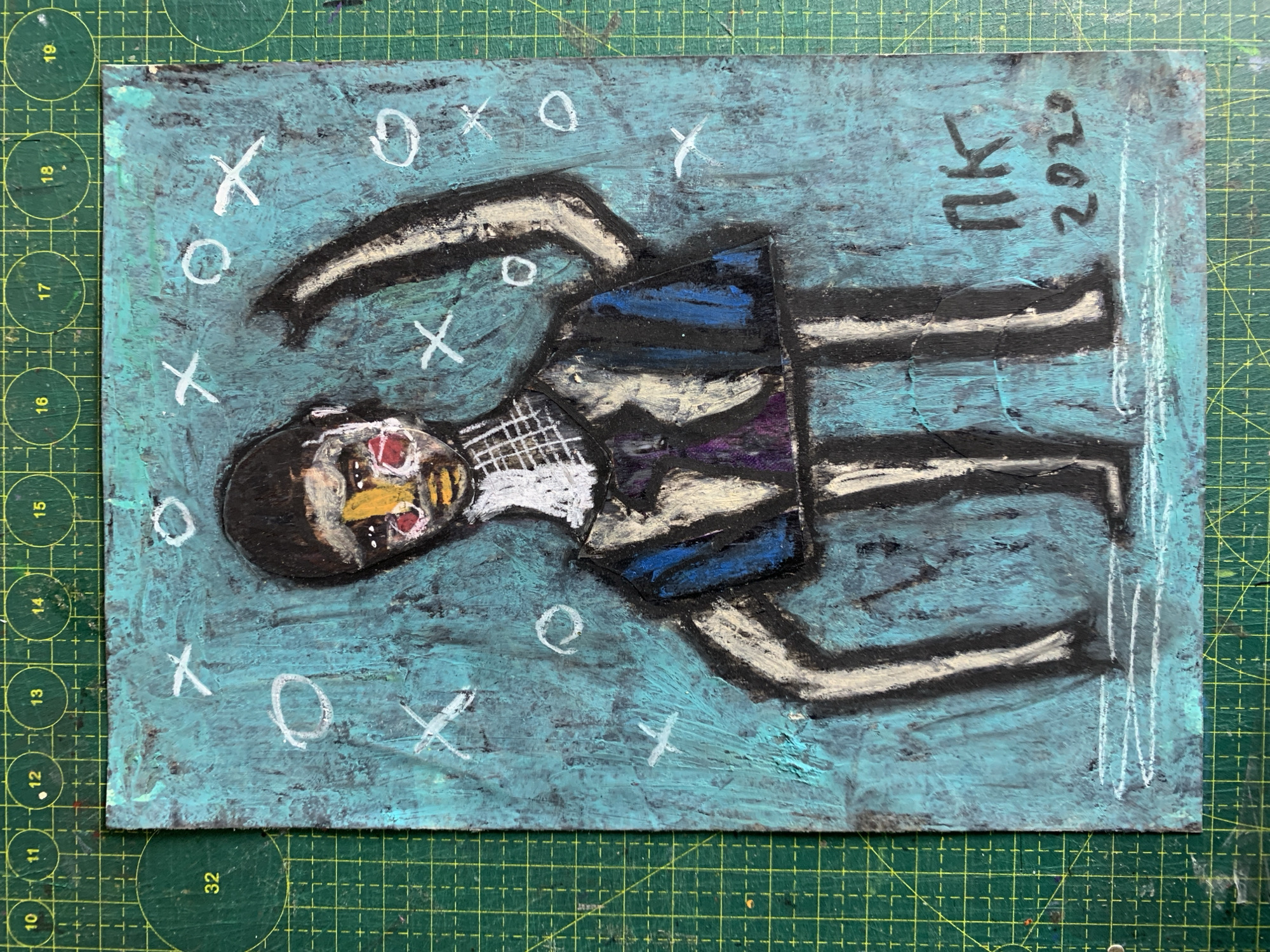 The image shows a piece of art with a dark green cutting mat as the background. The artwork itself is on a textured, teal-colored surface and depicts a stylized figure with a dark head of hair, yellow and red facial features, and a body consisting of a blue and black garment with a purple accent. The figure has multiple limbs, some of which are raised. White "x" and "o" shapes are scattered across the teal background. In the bottom right corner of the artwork, the initials "PK" and the year "2020" are visible. The artwork is positioned on the cutting mat, which has visible grid lines and circular markings. There is no indication of any specific location in Sergeevo, Russia, within the image itself.