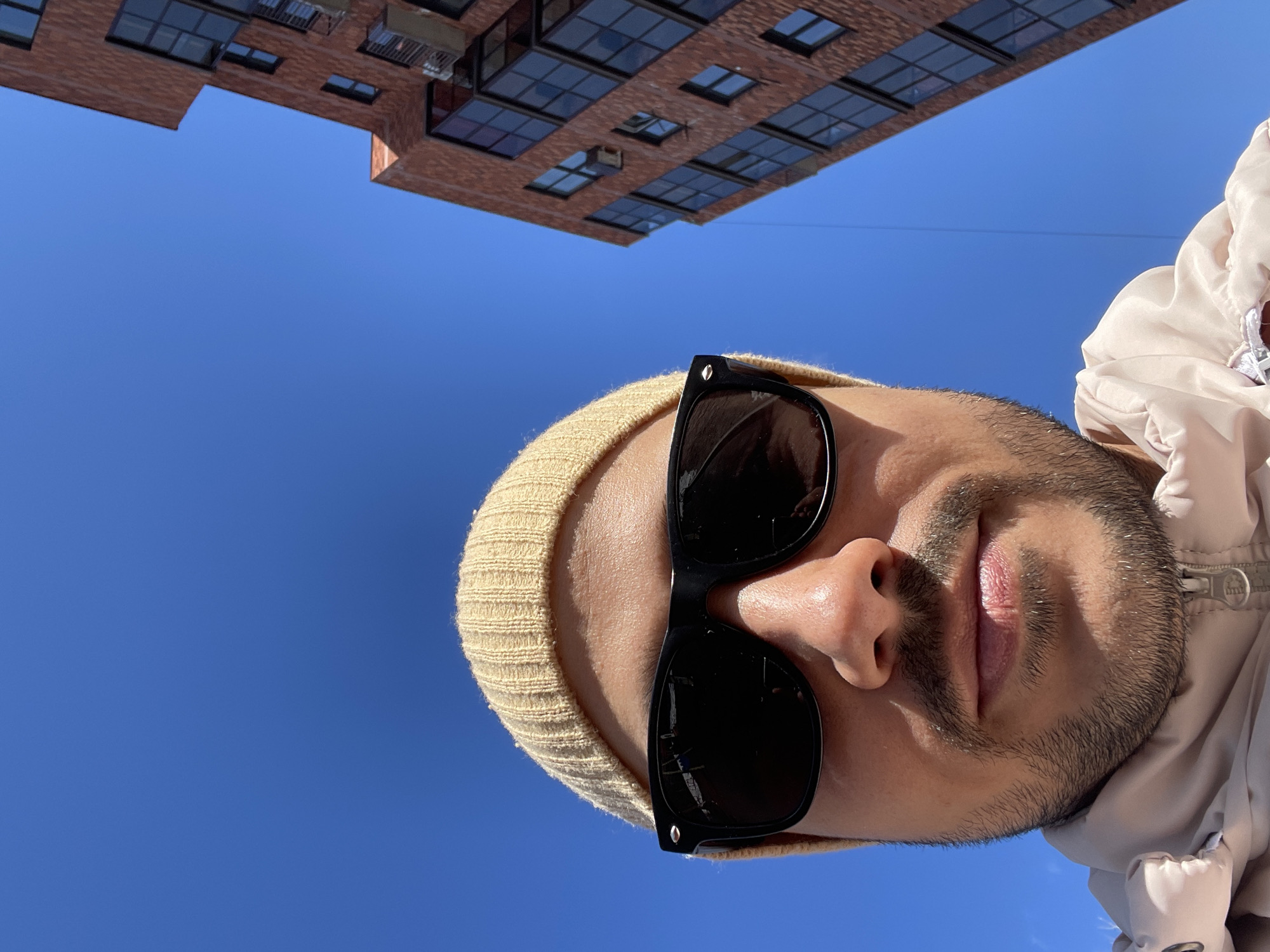 A low-angle selfie captures a person wearing a tan beanie and black sunglasses, with a glimpse of a beige jacket, set against a vivid blue sky. In the upper portion of the frame, a brick building with numerous windows is visible. This image was taken in Izhevsk, Russia.