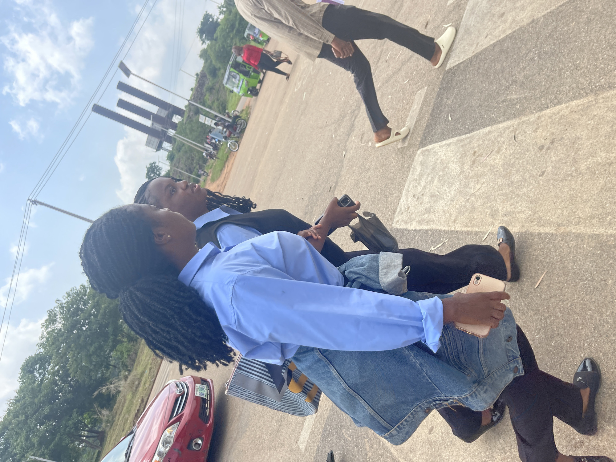 The image shows a group of people on a pedestrian crossing in Jos, Nigeria. Two women are in the foreground, sitting. The woman on the left is wearing a light blue collared shirt and jeans, with her hair in long braids. She is holding a phone in her right hand. The woman on the right is wearing a black outfit and is partially visible. Behind them, a man in a striped shirt and black pants is walking across the street. Further in the background, a bright green tuk-tuk is visible, along with several motorcycles and a red car. The scene appears to be outdoors during the day, with a cloudy sky. The ground is paved, with distinct white lines marking the pedestrian crossing. Trees and hills are visible in the distance. The overall atmosphere seems casual and ordinary, suggesting daily life in a Nigerian urban or semi-urban setting.