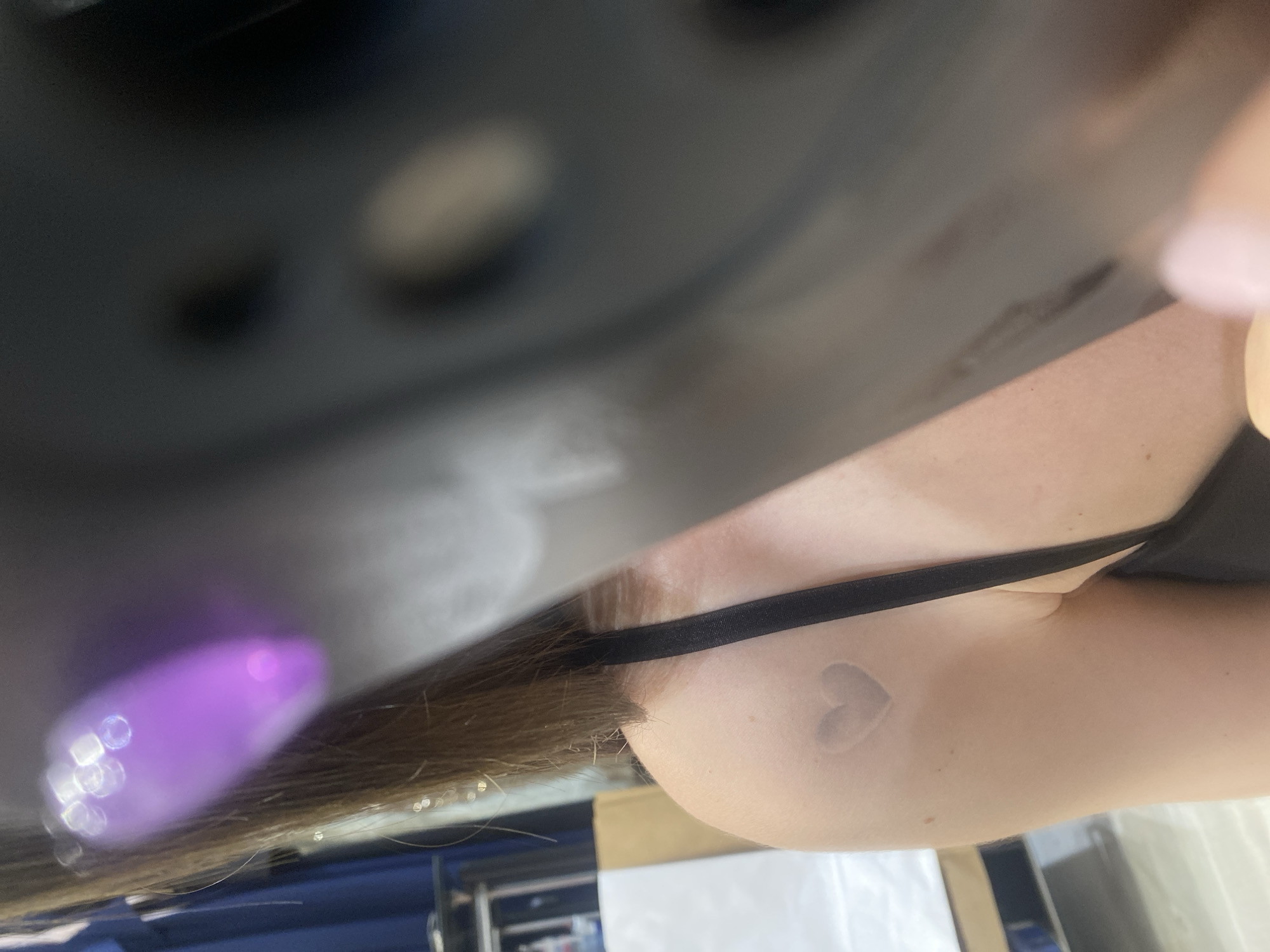 An out-of-focus, medium shot captures a portion of a person's upper back and shoulder. A faint, light-colored heart-shaped mark is visible on the person's left shoulder. A black bra strap runs diagonally across the person's upper back. The background is largely obscured by a dark, blurry object in the foreground, though hints of clothing and a beige surface are discernible. The lighting suggests an indoor setting. The image is not from a city.